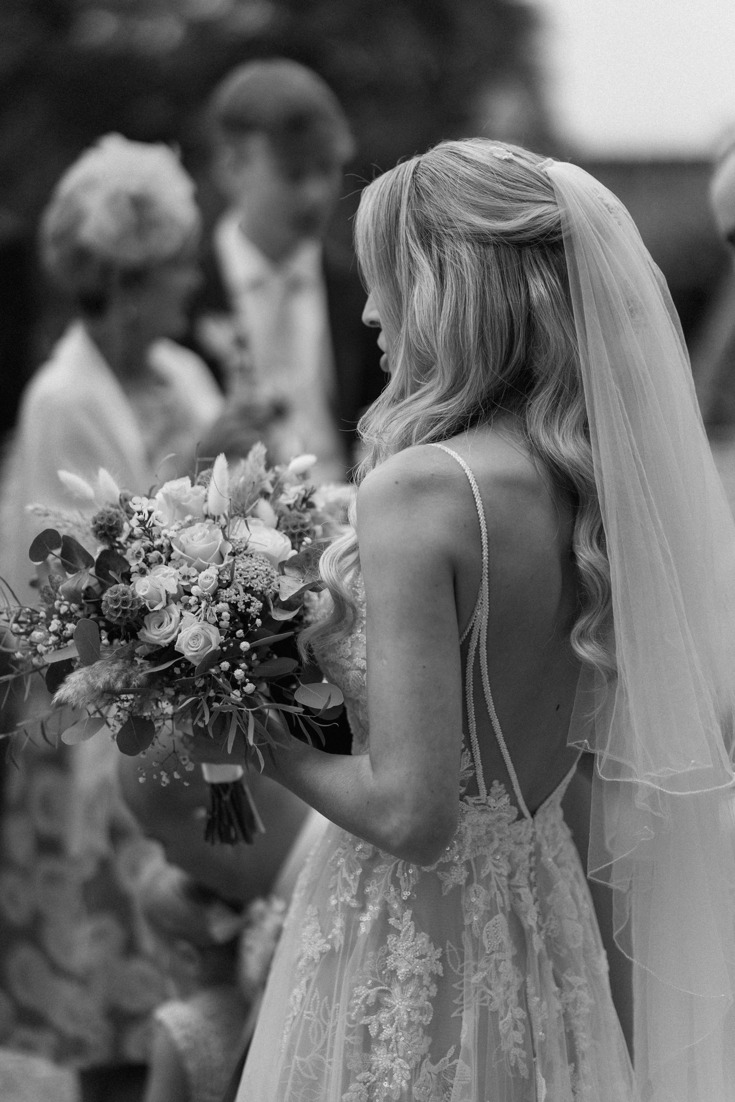 Elegant bride in sleeveless lace gown holds beautiful floral bouquet during outdoor ceremony in Warwickshire.