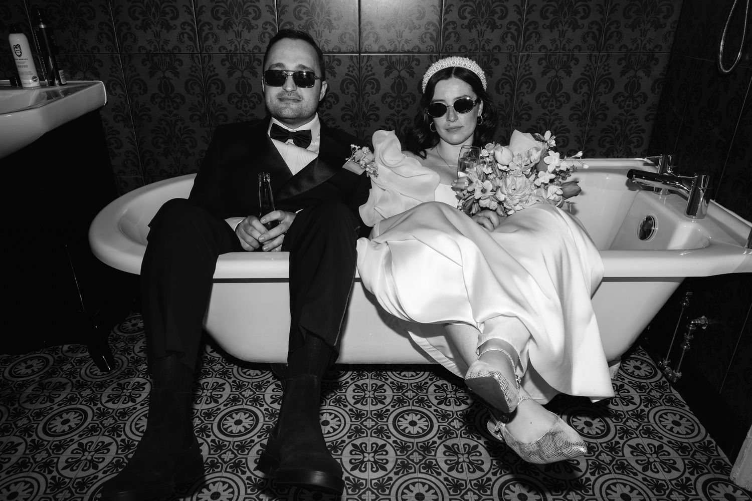Black and white photo of a couple in wedding attire wearing sunglasses, sitting in a bathtub on patterned tile floor.