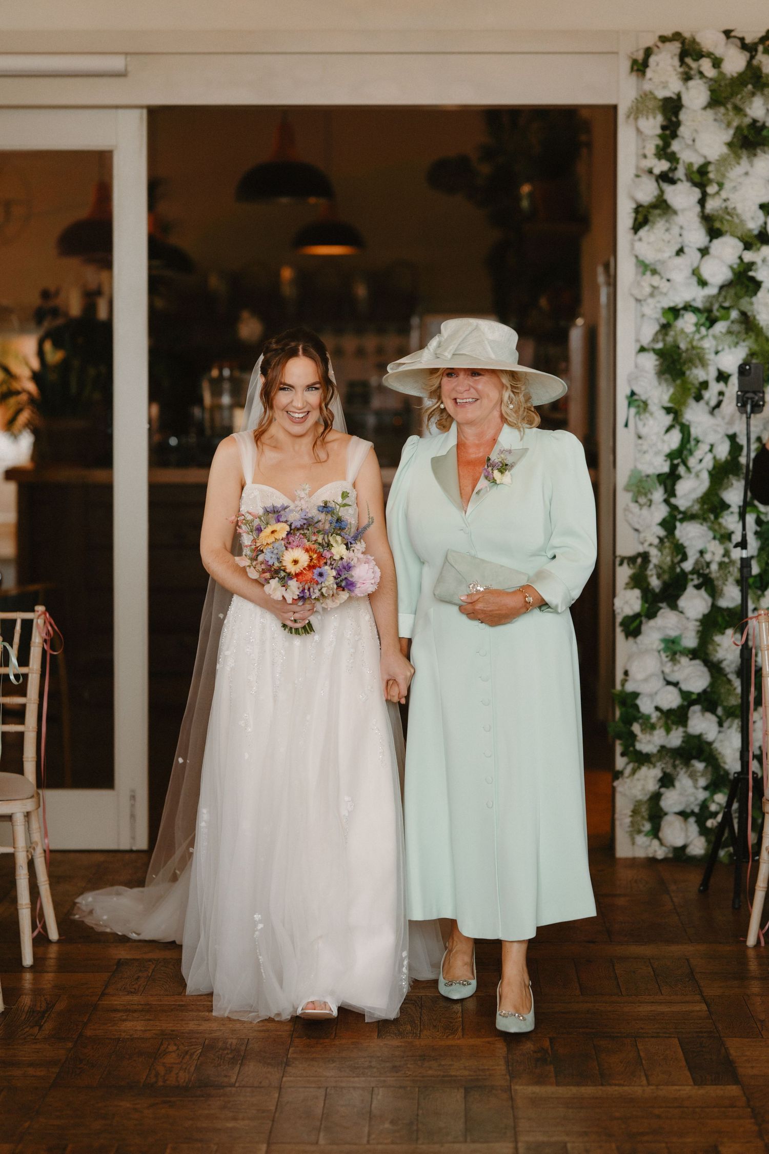 Bride in white tulle gown holding colorful bouquet walks with older woman in mint green outfit.