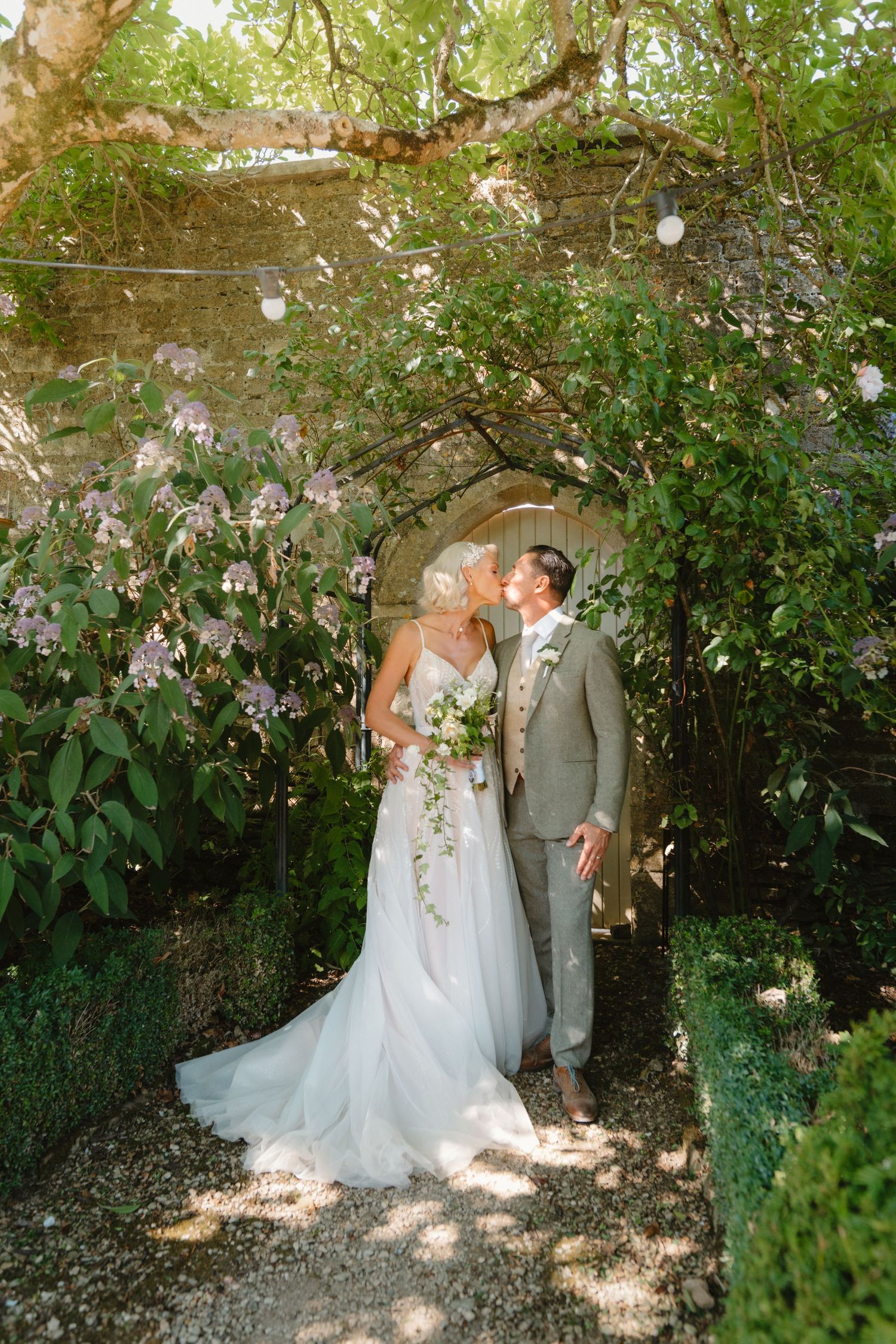 Bride and groom sharing a kiss under a lush floral arch with climbing roses at a garden wedding venue.