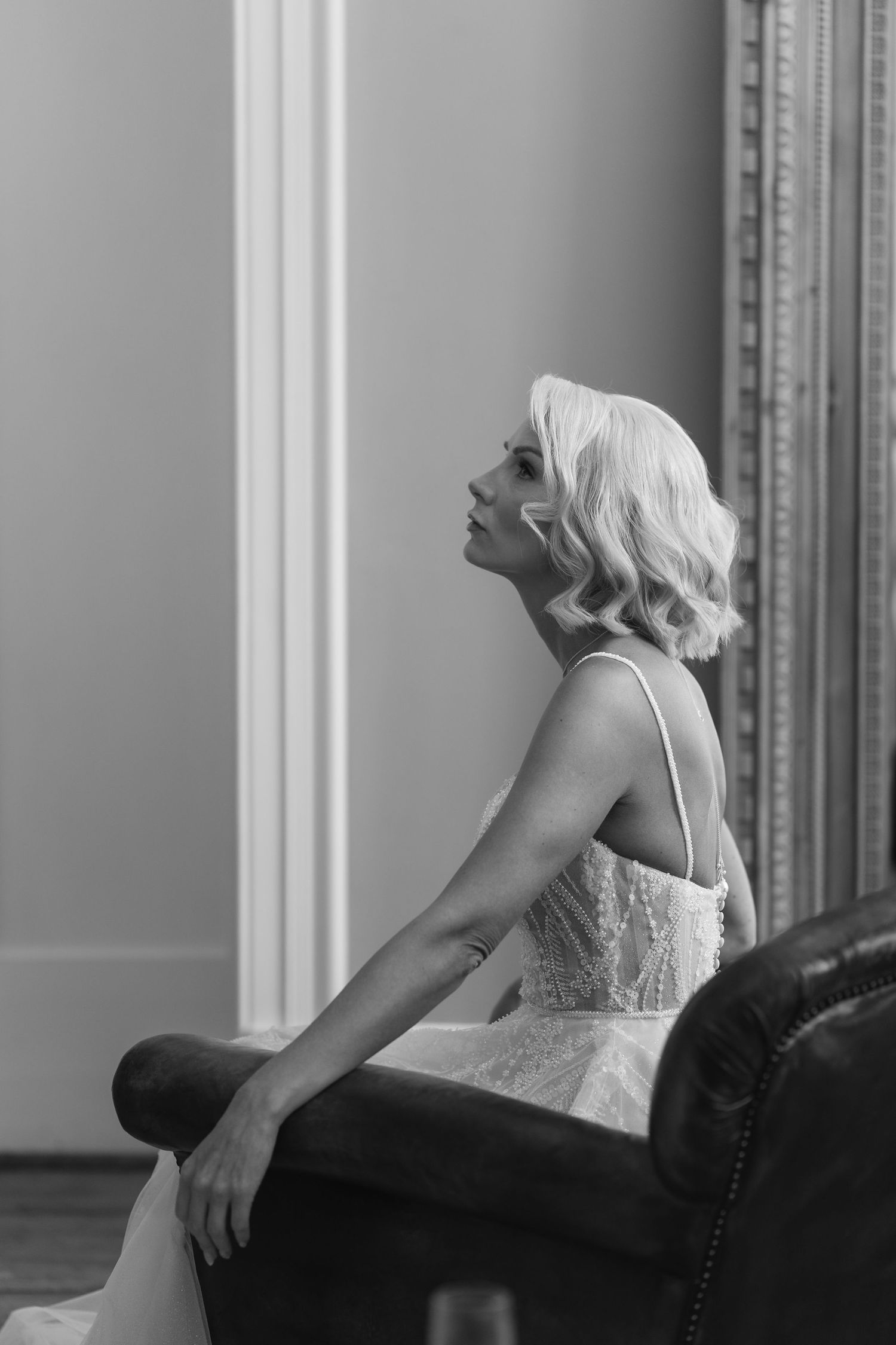 Black and white photo of an elegant bride in a lace gown sitting on a leather sofa, gazing upward in a classic interior.