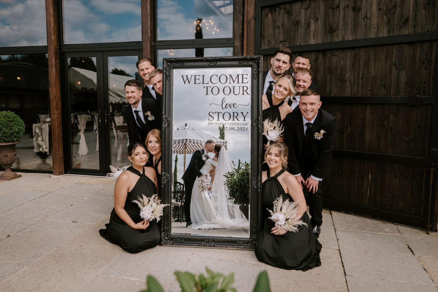 Wedding party poses around a welcome sign at a rustic venue with wooden backdrop.