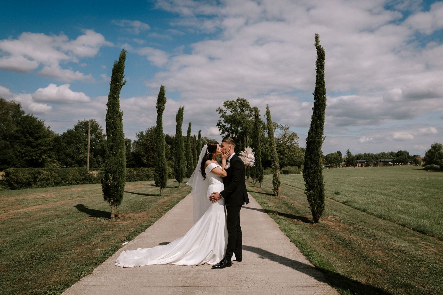 Couple shares romantic moment on path lined with tall cypress trees on wedding day.