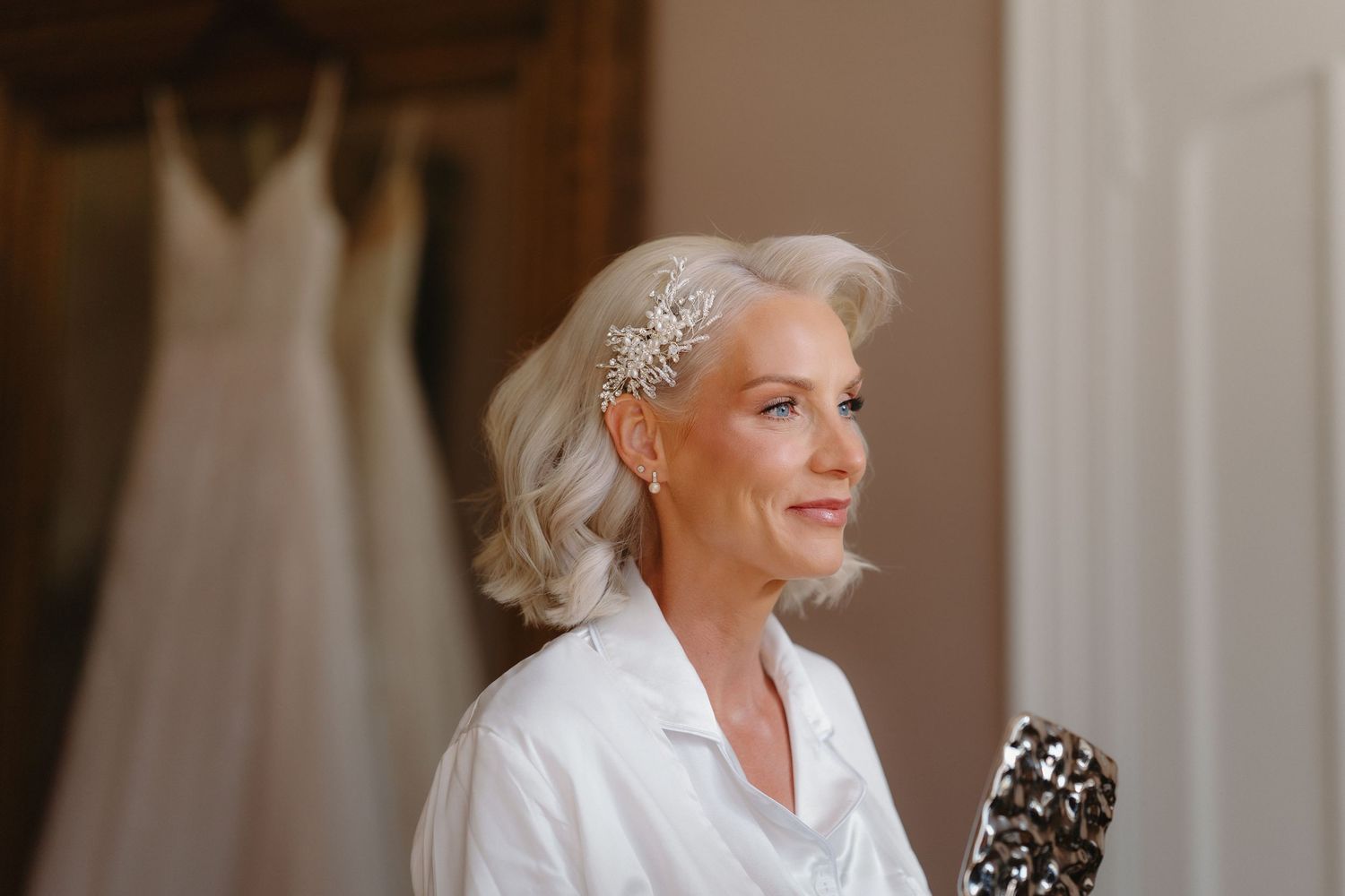 Elegant bride with silver hair wearing a white robe and crystal hair accessory, with wedding dress hanging in background.