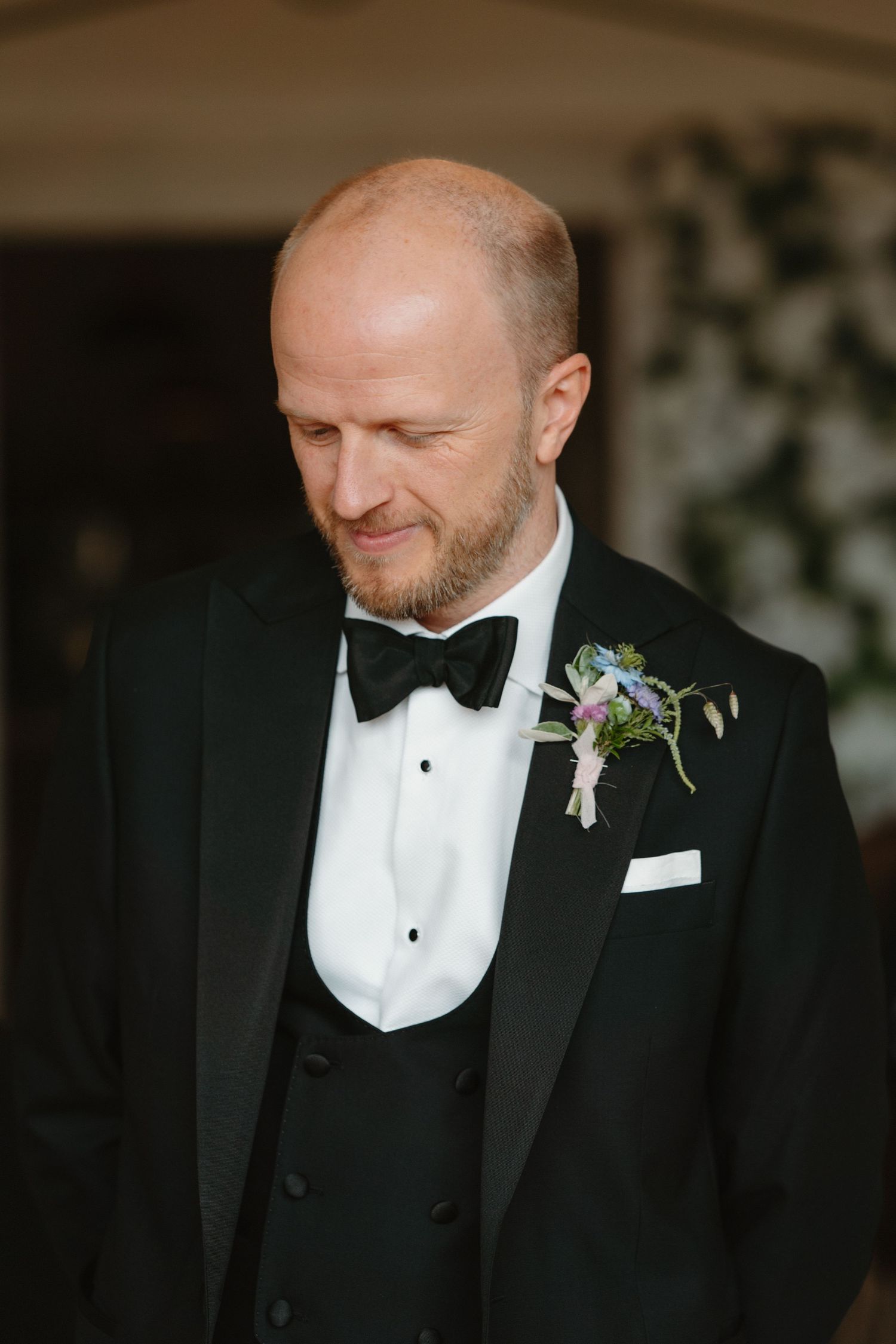 Groom in black tuxedo with bow tie and floral boutonniere smiling downward at wedding ceremony.