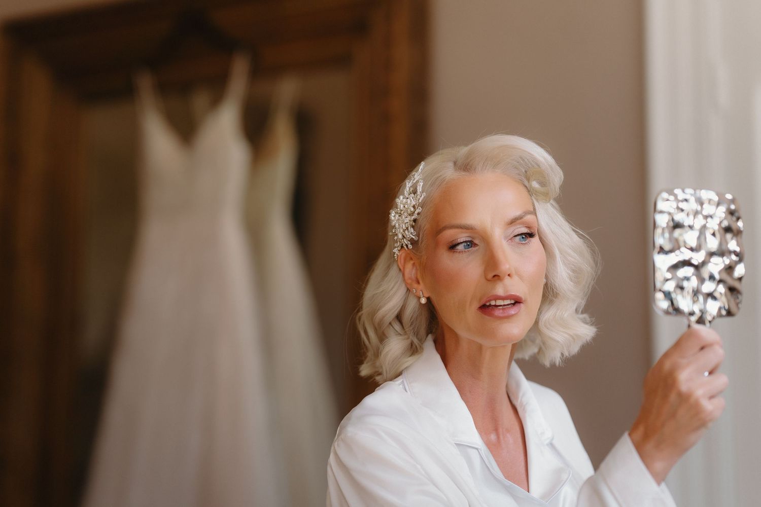 Elegant older bride with silver hair checks makeup in ornate mirror, wearing white robe with wedding dress hanging in background.