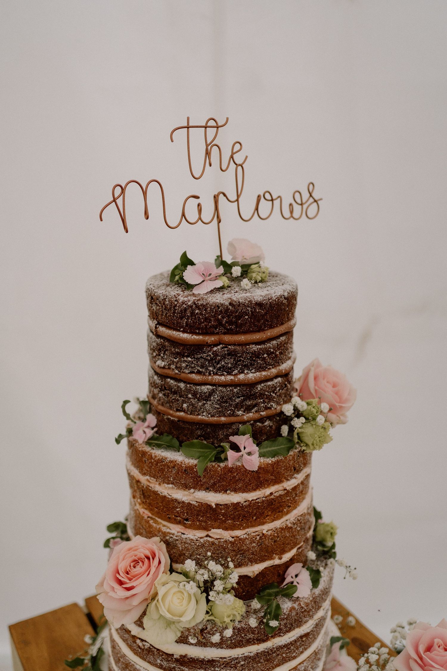A rustic naked wedding cake decorated with pink roses and greenery, topped with 'The Marlows' script topper.