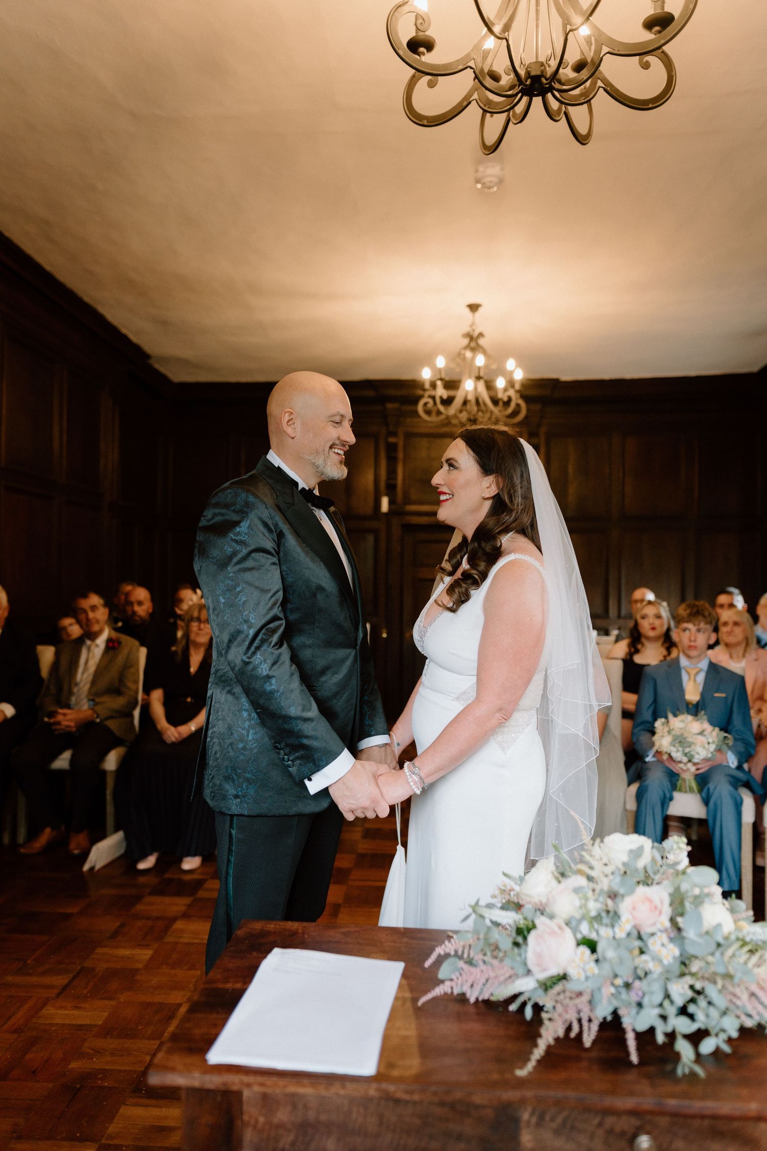 A wedding ceremony takes place in an elegant wood-panelled room with chandeliers and gathered guests at St Johns House in Warwick.