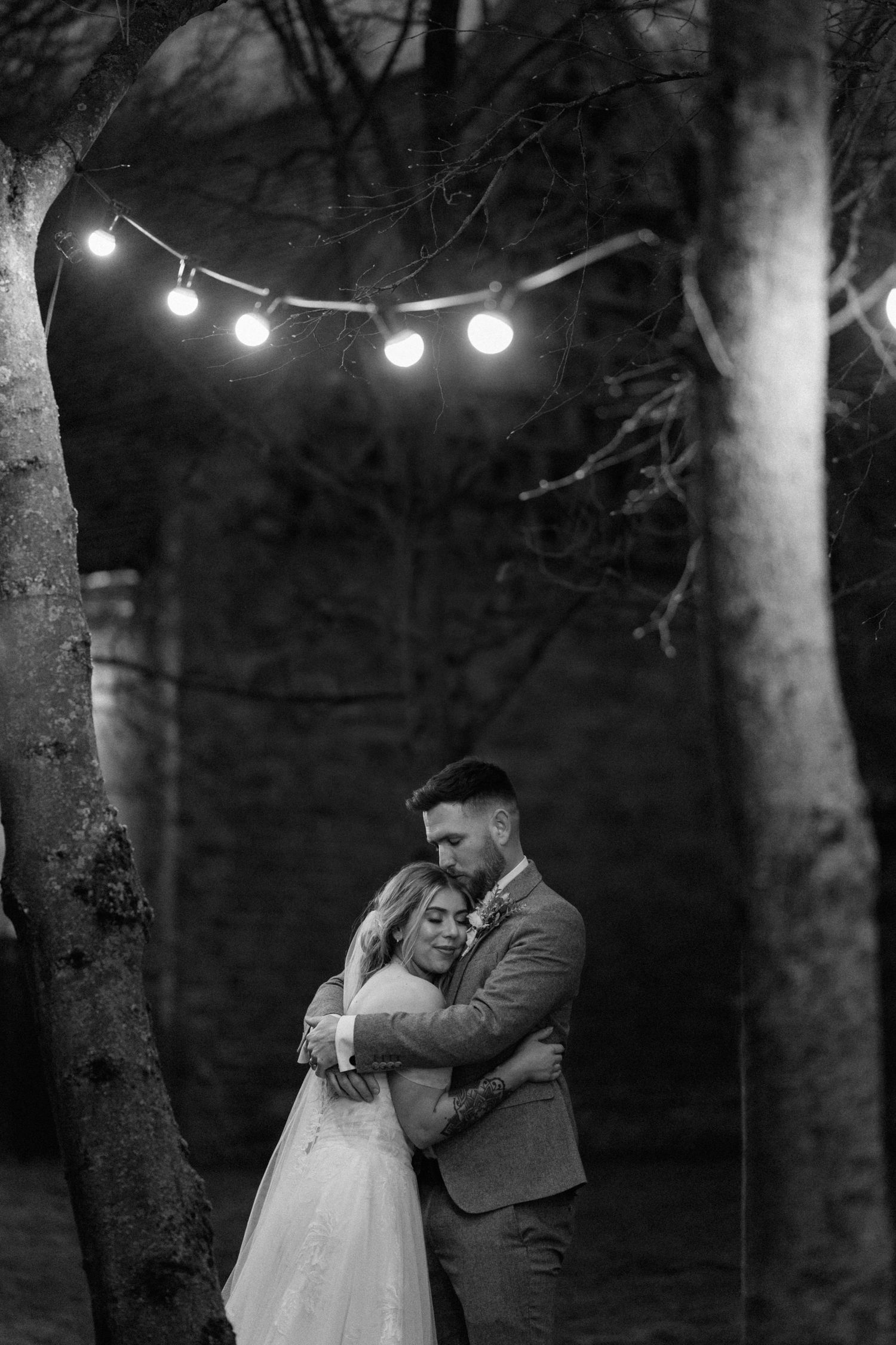 A romantic moment between newlyweds under string lights in a dark forest setting at Shustoke Barn.
