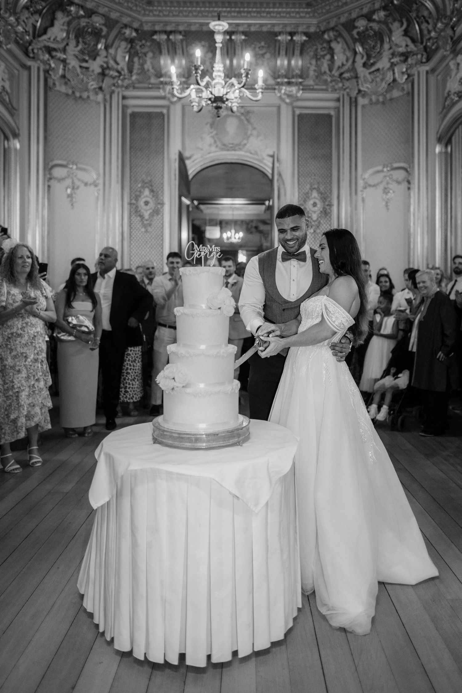 A wedding reception in an elegant ballroom with a tall white cake and crystal chandelier at Harlaxton Manor.