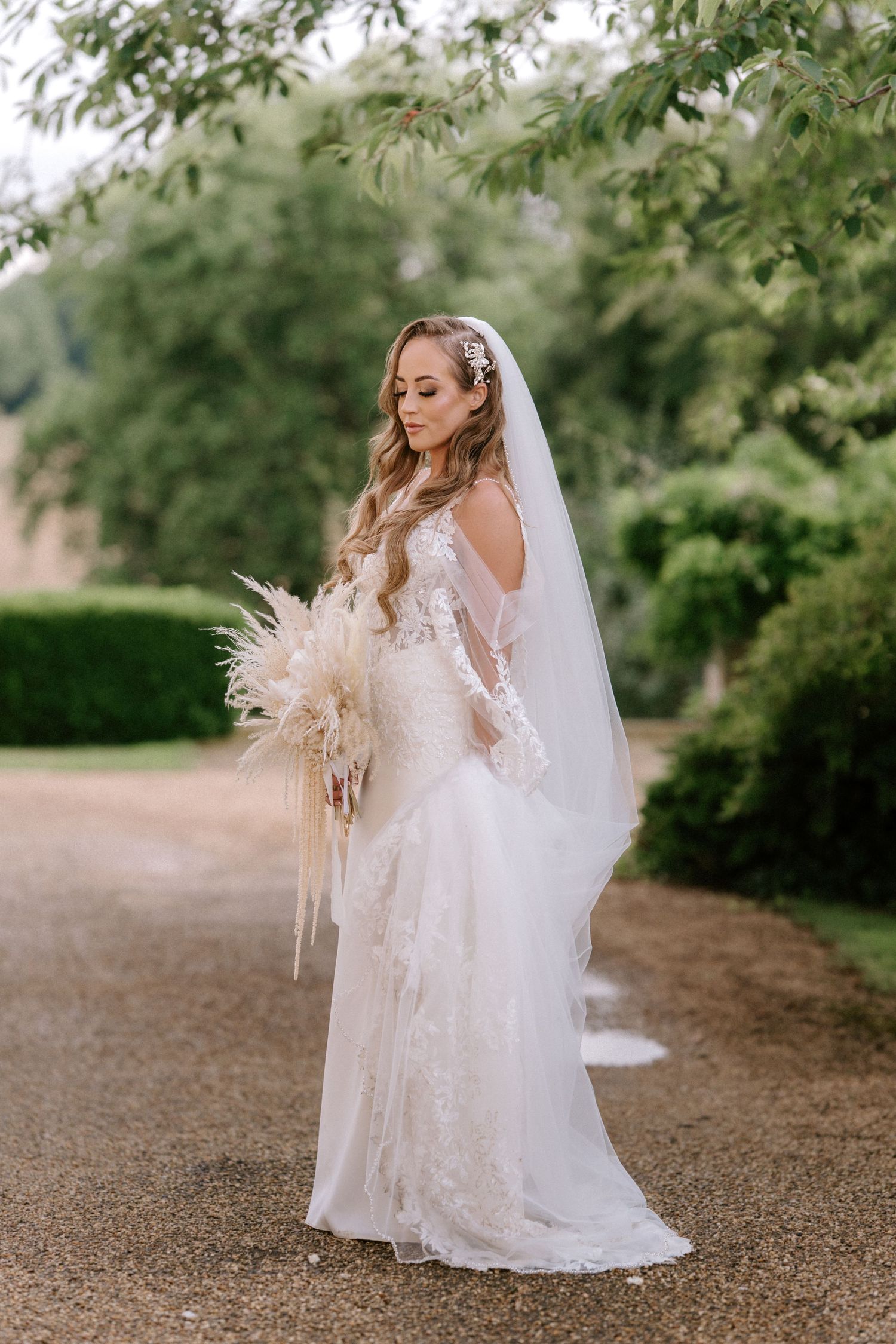 A bride in a lace wedding gown holds a dried pampas grass bouquet. Wedding photography at Rushton Hall. Northamptonshire Wedding photograph