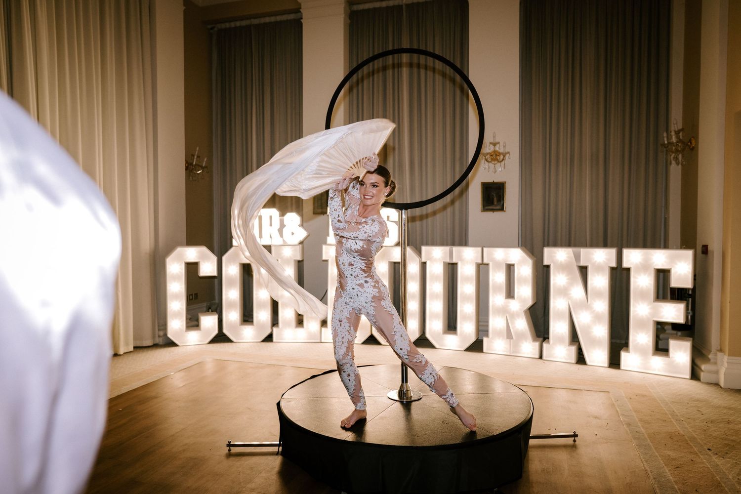A performer in a sparkly outfit spins a hula hoop on an illuminated circular stage with 'COLBOURNE' text display.