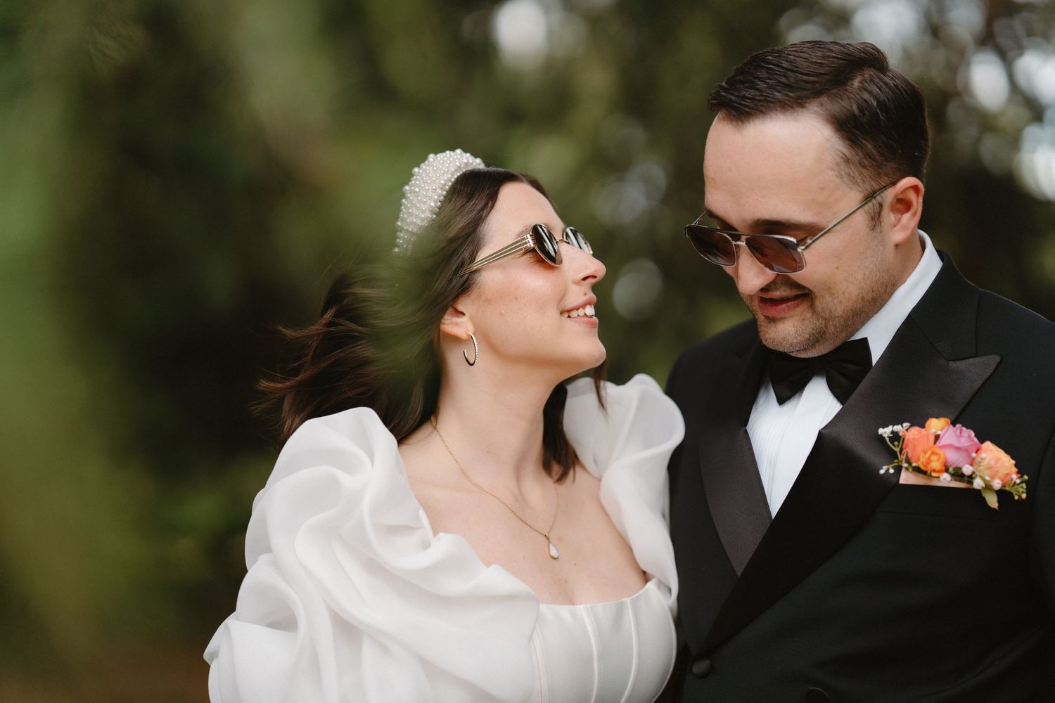 Bride and groom in sunglasses smiling at each other outdoors, groom in tuxedo with floral boutonniere.