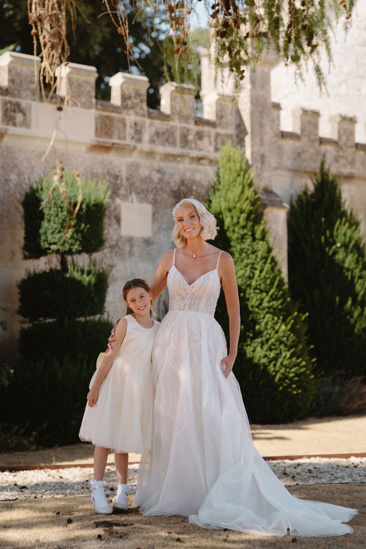 Smiling bride in lace wedding gown poses with young flower girl in white dress at a castle garden.