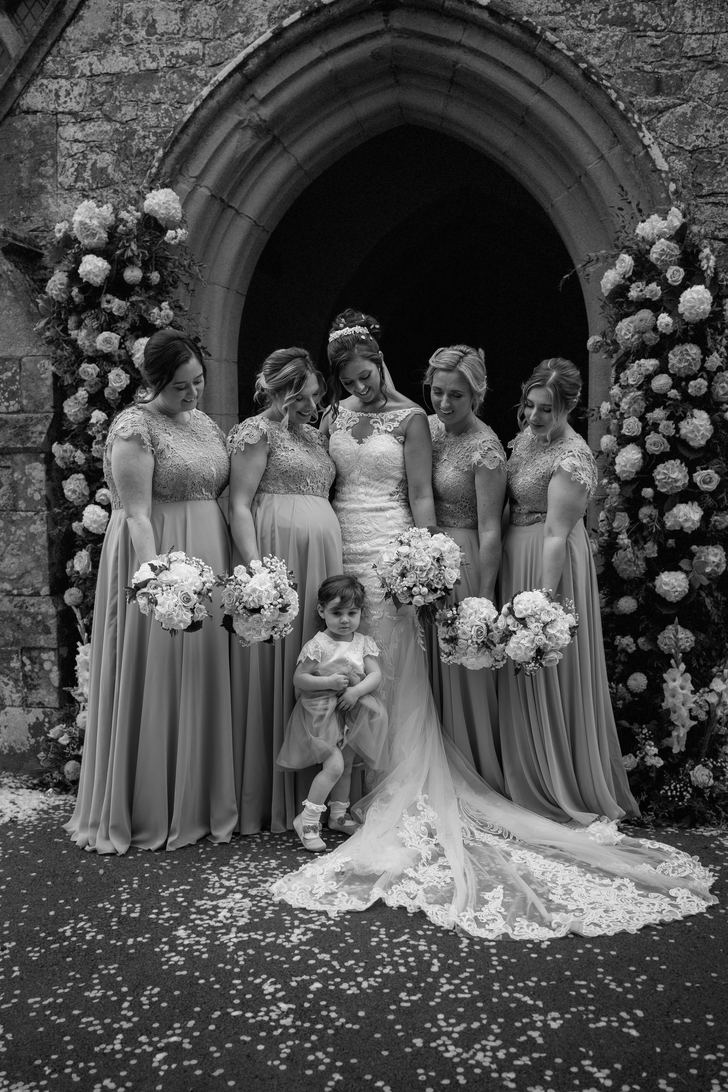 Elegant black and white wedding photo of bridal party gathered under stone church archway with floral decorations.