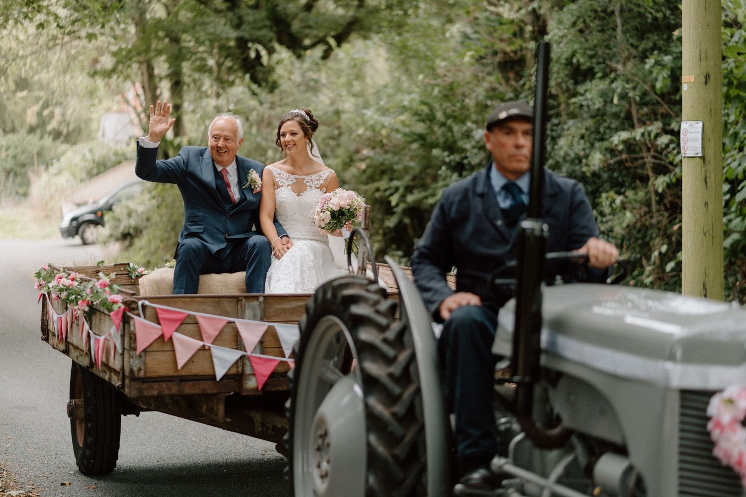 A couple rides in a decorated vintage tractor trailer along a tree-lined road during a countryside celebration.