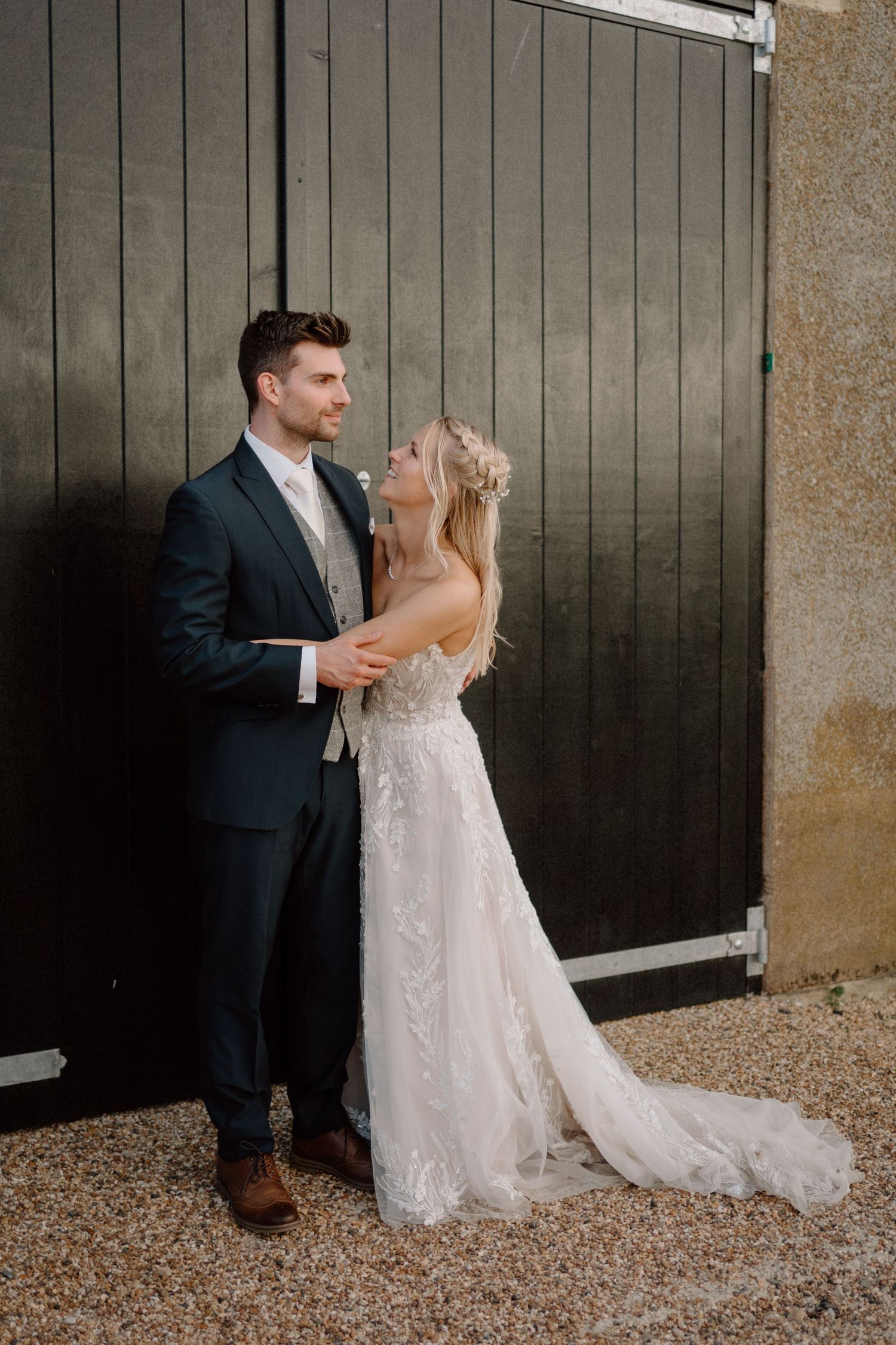 A bride in a lace wedding gown and groom in navy suit share an intimate moment against a rustic barn door at Warwick House.