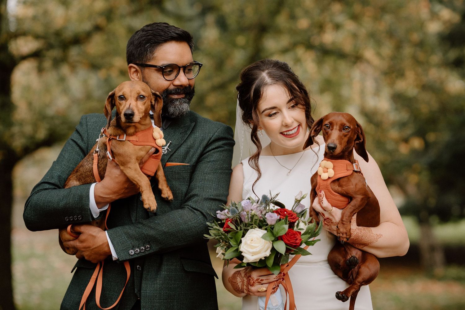 A wedding couple poses outdoors while holding two small brown dachshund dogs and a floral bouquet at The Glasshouse in Leamington Spa.