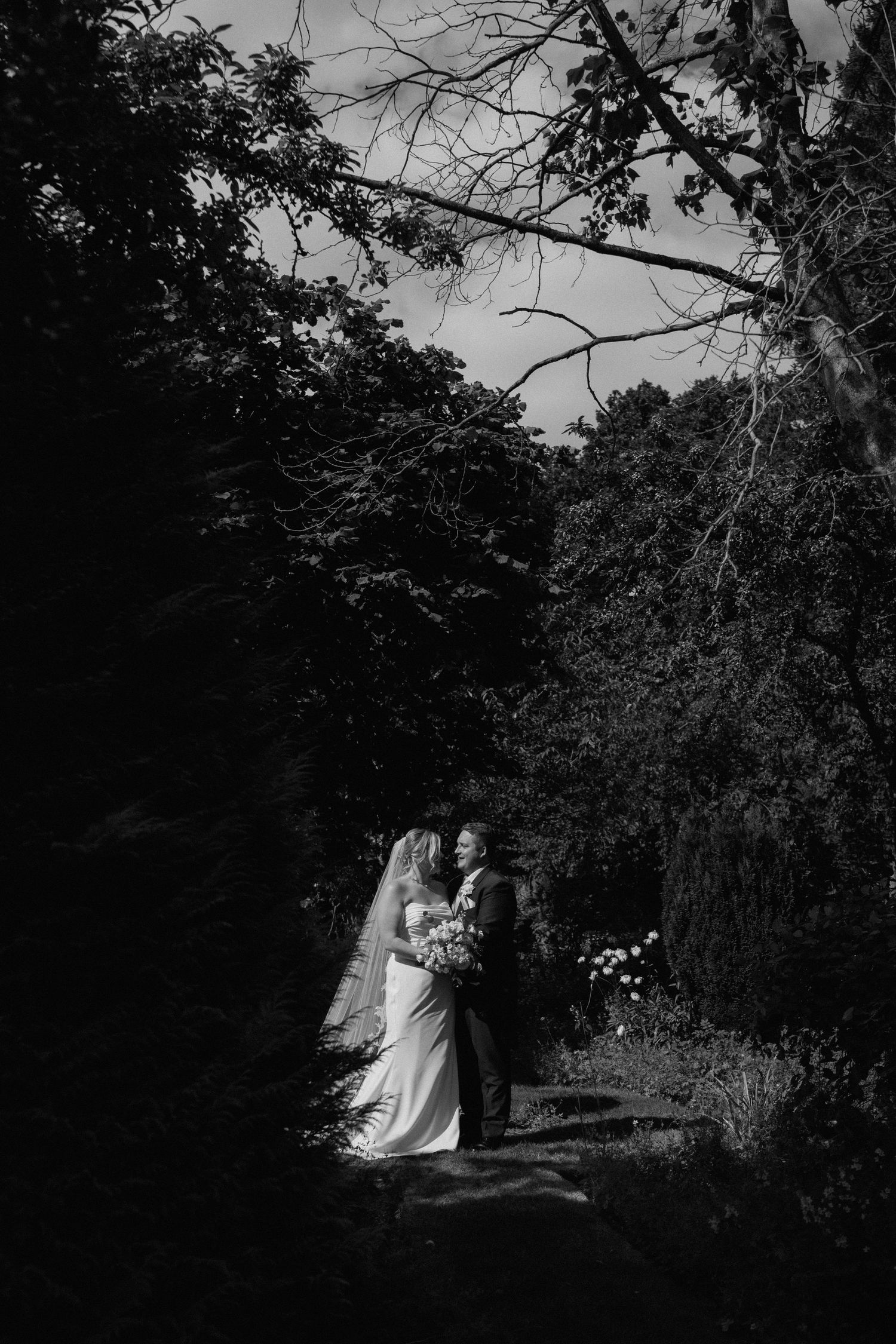 A romantic black and white wedding photo of a couple standing on a garden path surrounded by trees at Nuthurst Grange.
