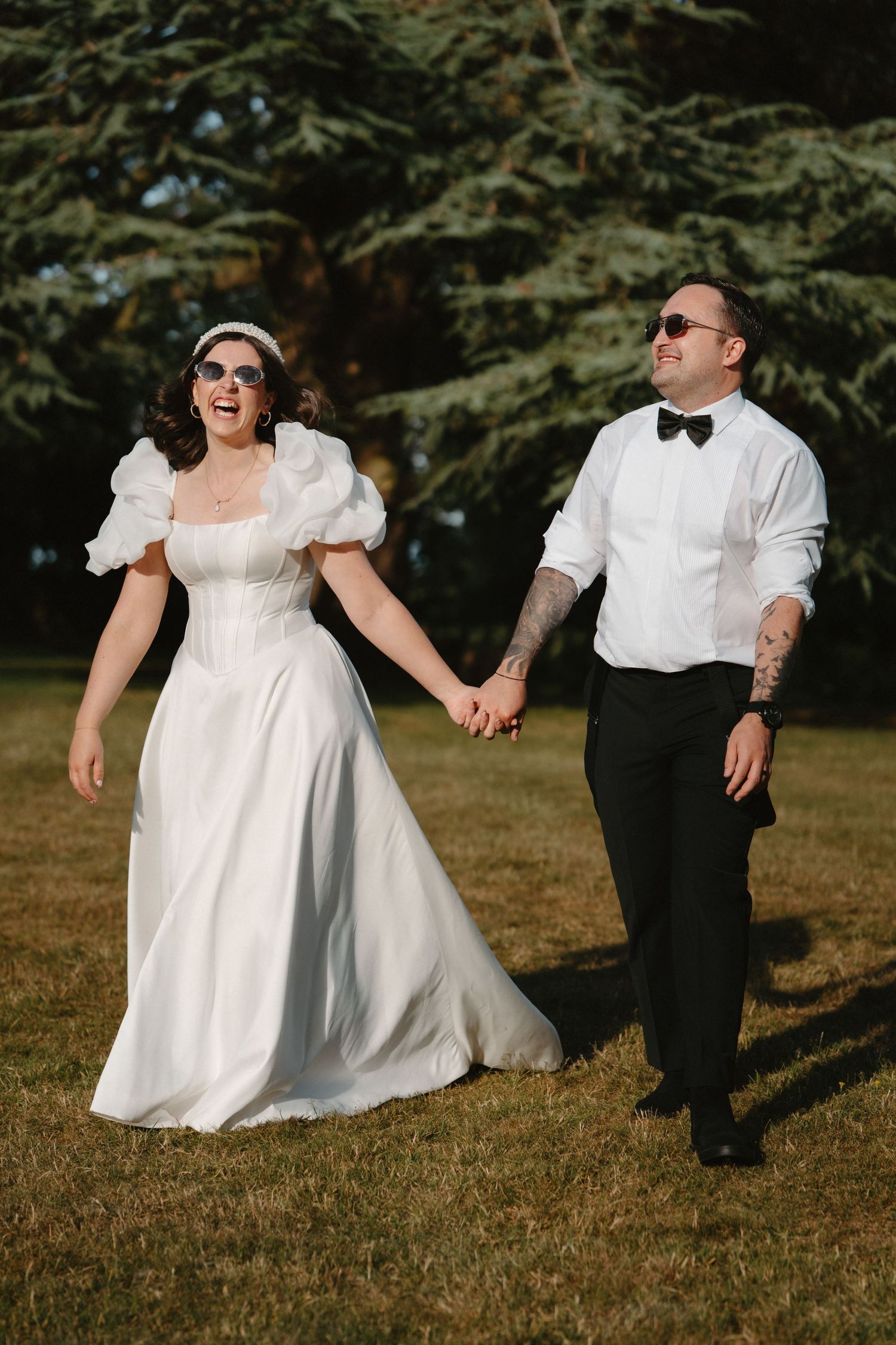 A joyful bride in a white puff-sleeve gown and groom in bow tie hold hands walking outdoors on their wedding day.