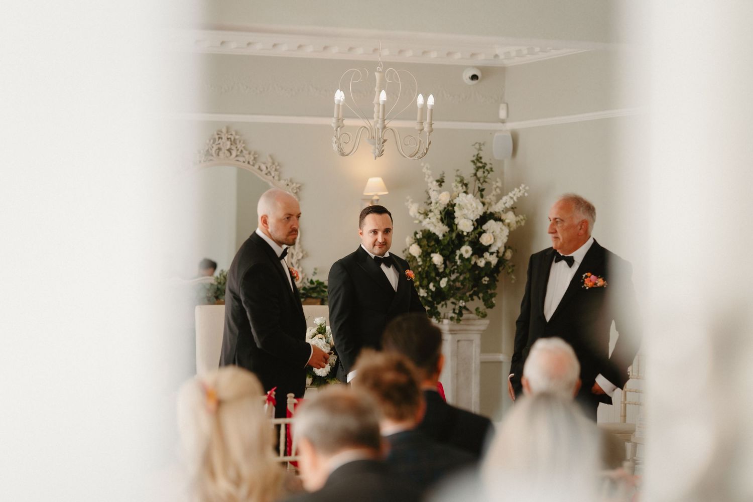 Three men stand at a wedding ceremony altar in an elegant white room with chandelier and floral arrangements.