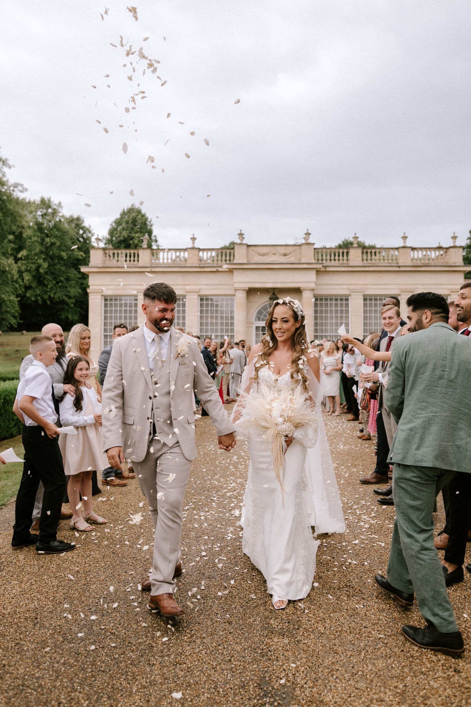 Newlyweds walk down path as wedding guests shower them with confetti outside an elegant stone venue.
