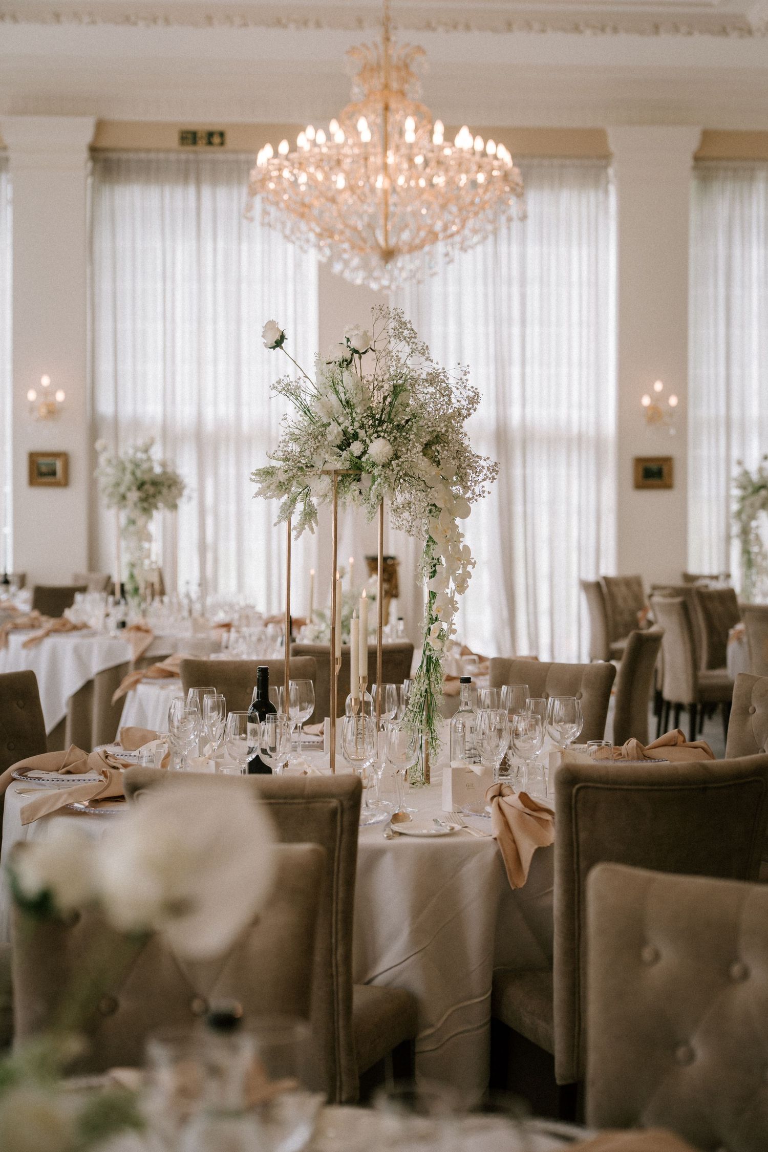 Luxurious wedding reception room with crystal chandelier and tall white floral centerpieces.