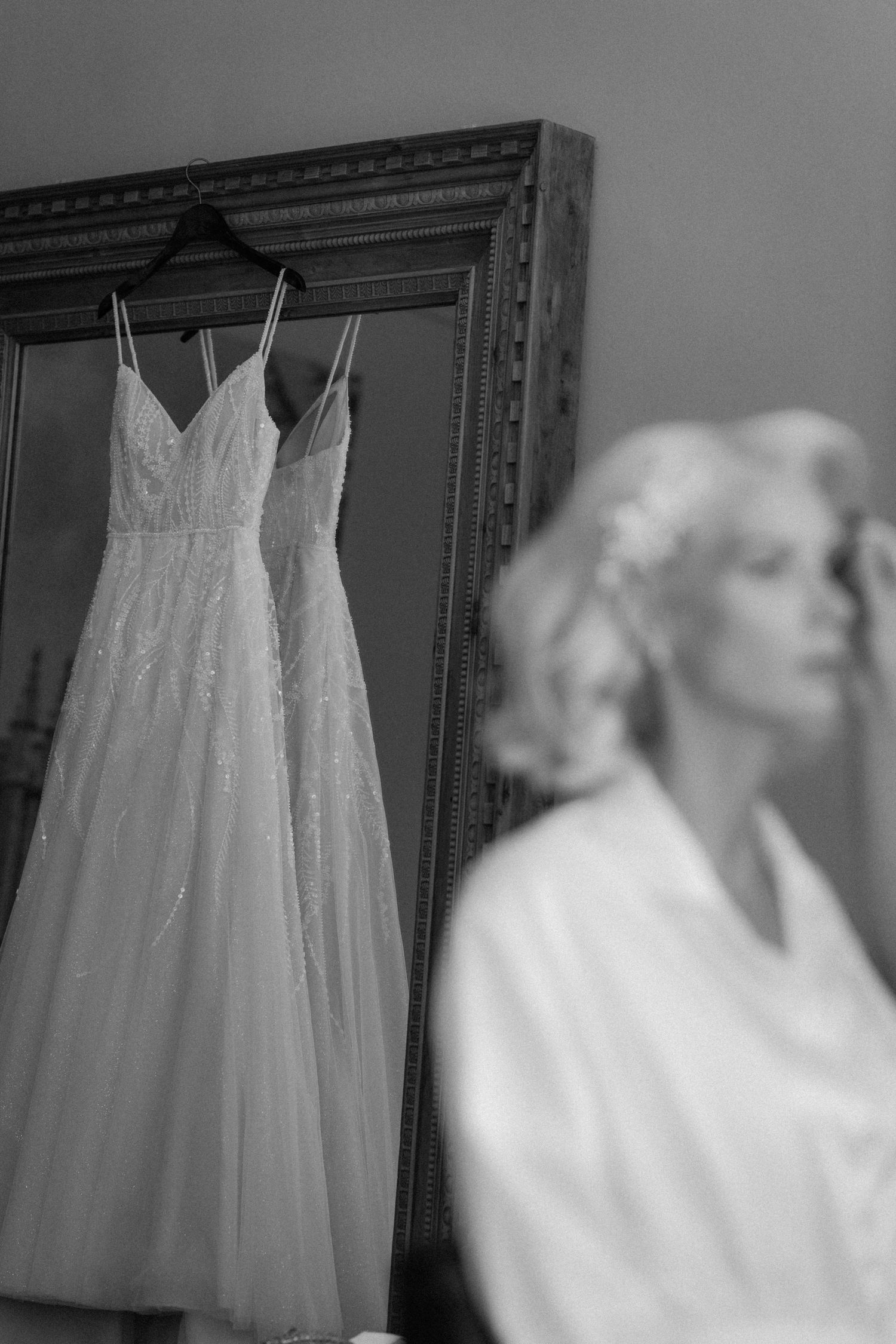 Black and white photo of a bride in a robe gazing at her wedding gown hanging in an ornate mirror.