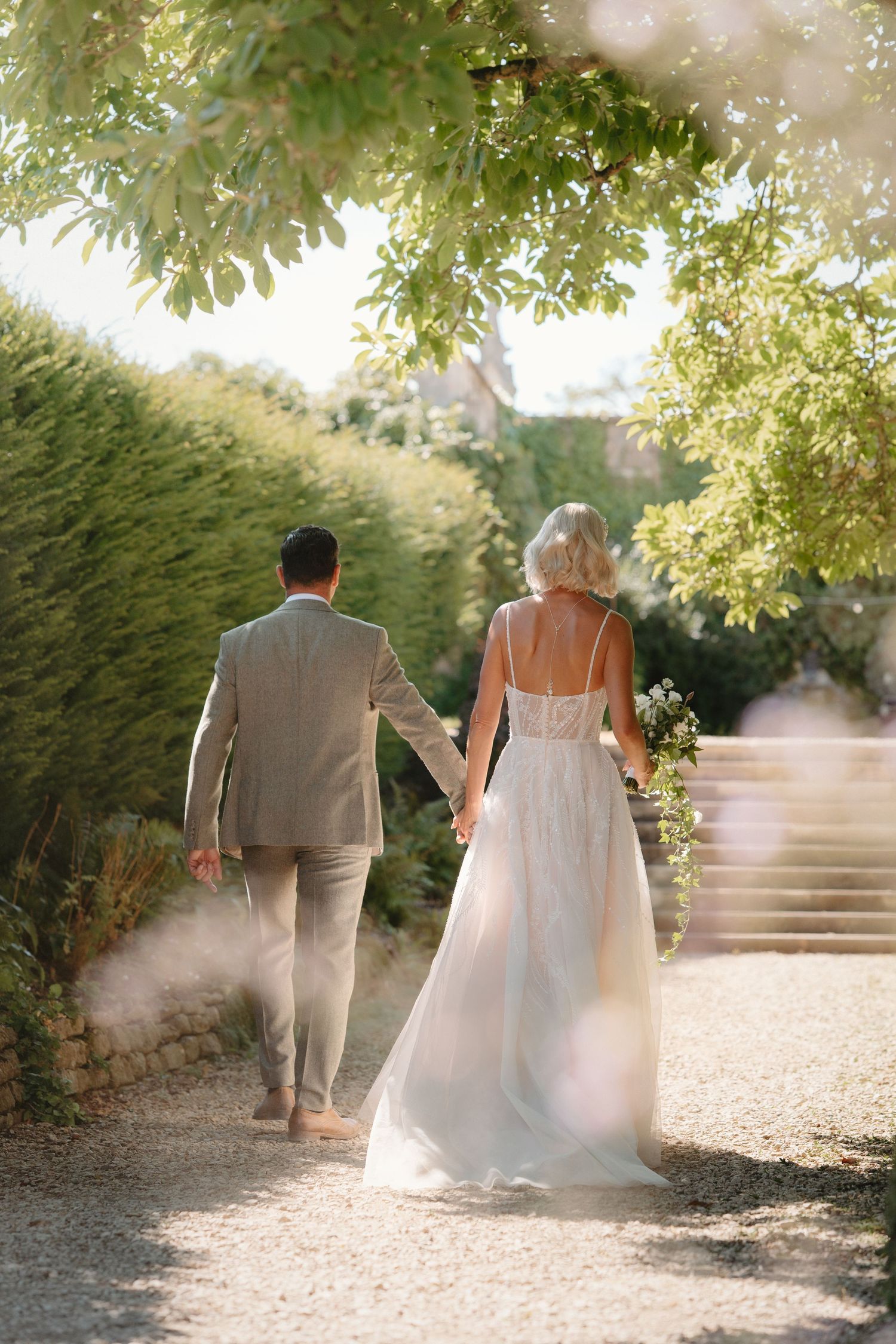 Bride and groom walking hand in hand down a sunlit garden path, bride in a flowing white gown holding a bouquet.