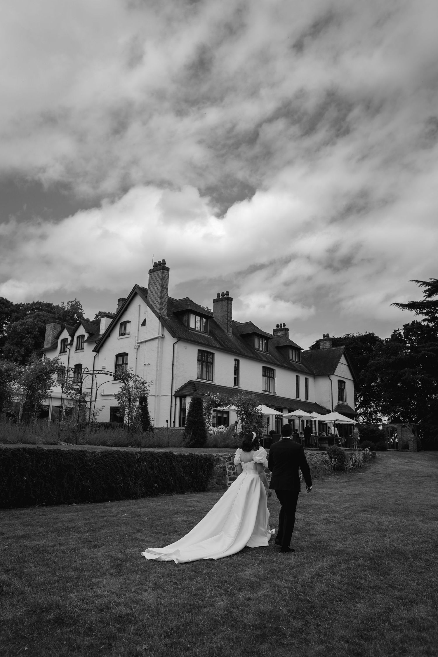 Black and white photo of bride and groom walking toward elegant manor house venue under dramatic cloudy sky.