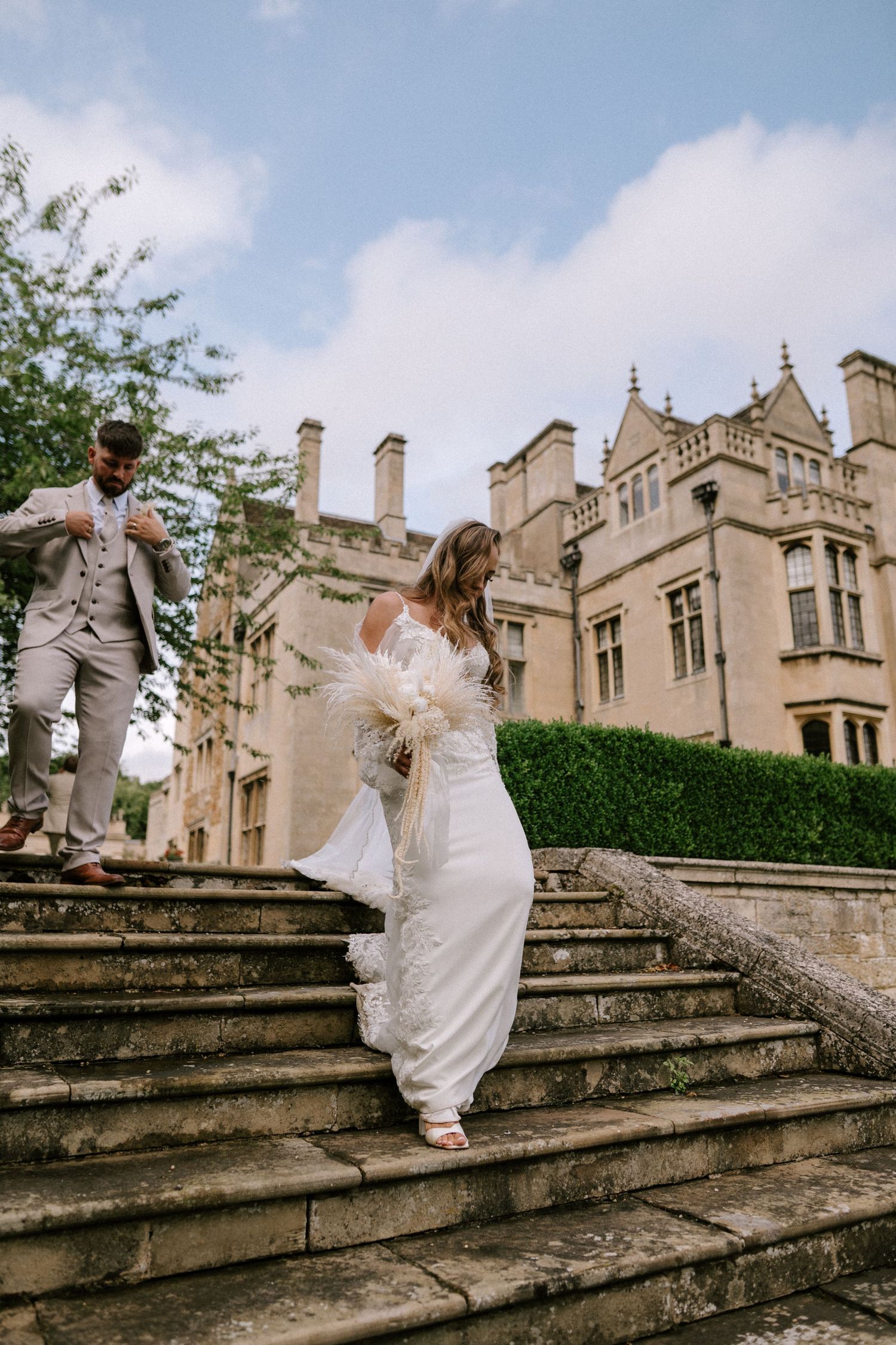 A couple ascends grand stone steps outside a Gothic-style mansion on their wedding day.
