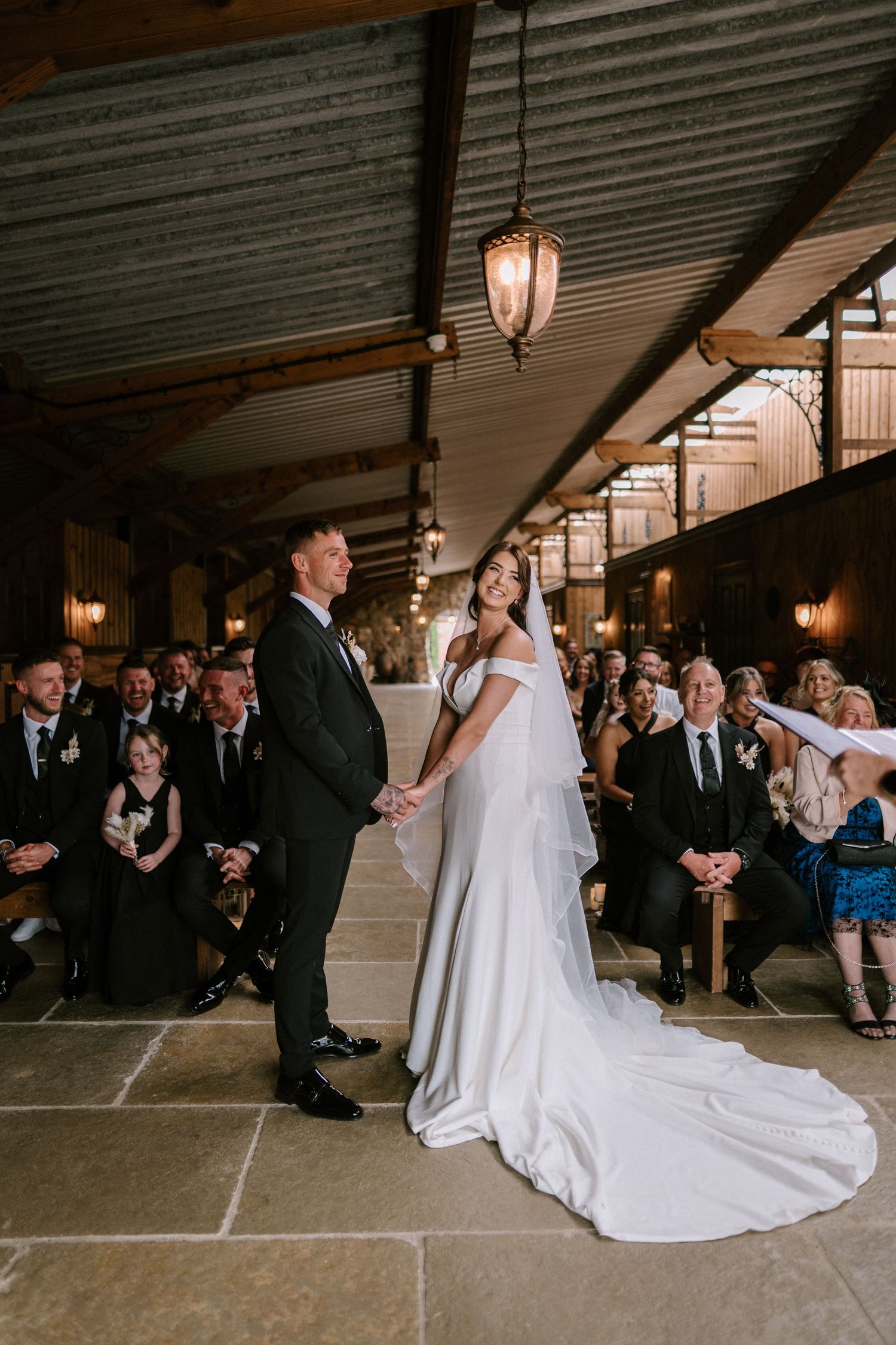 A wedding ceremony takes place in an industrial-style venue with exposed beams and hanging lanterns.
