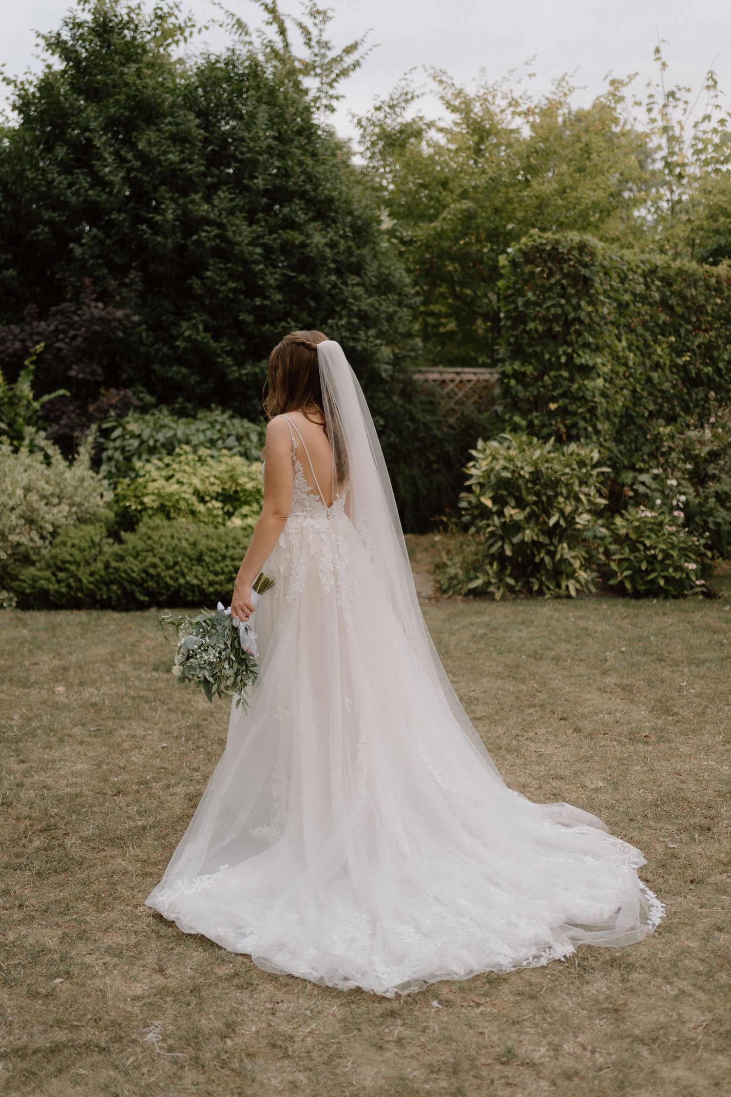 A bride in a flowing white lace wedding dress with long veil stands in a garden holding a bouquet at Warwick House.
