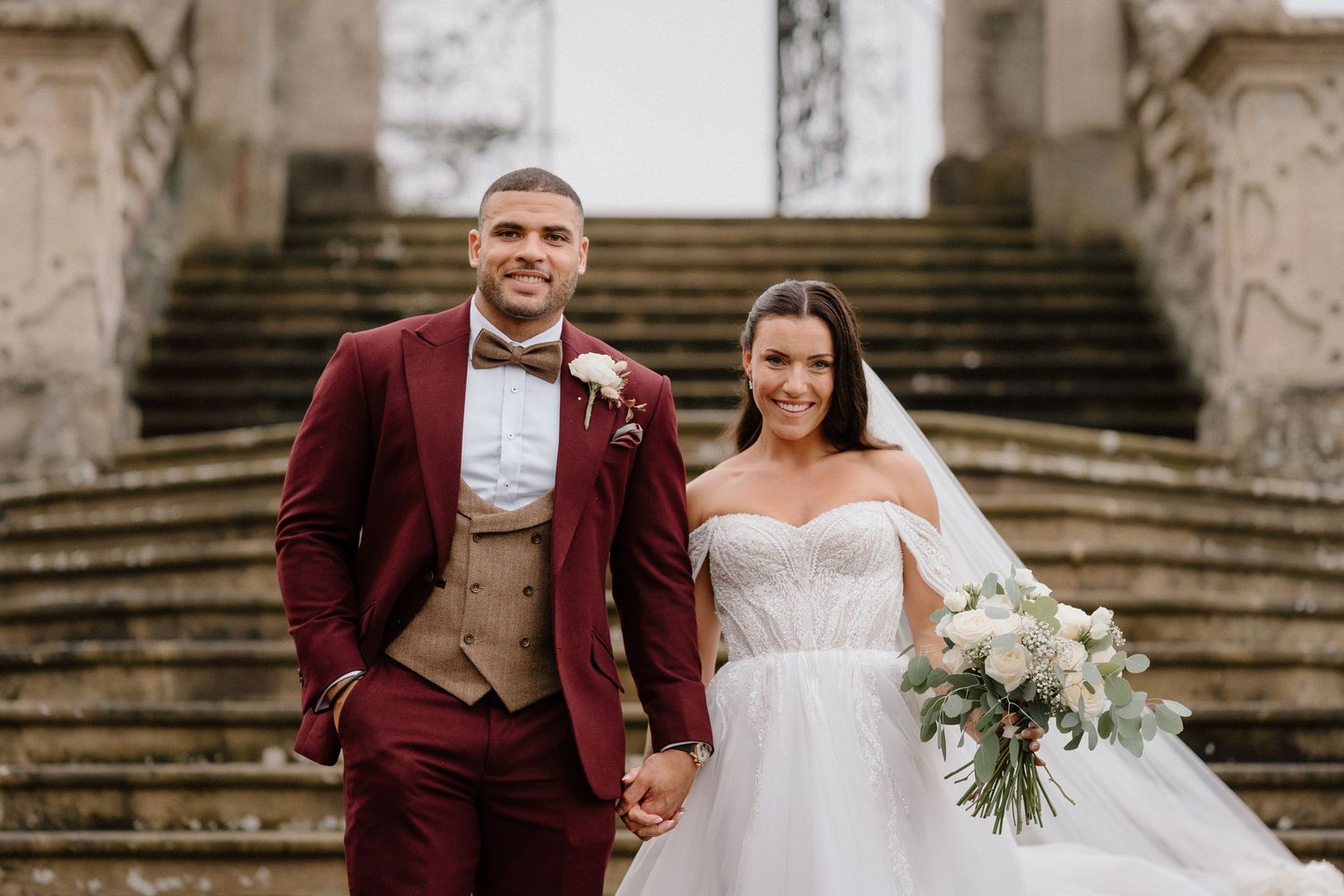 A wedding couple poses on stone steps, with groom in burgundy suit and bride in white gown with floral bouquet at Harlaxton Manor.