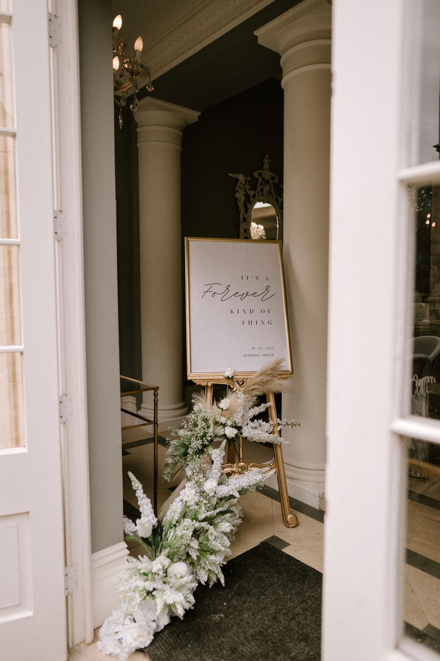 Elegant wedding welcome sign display with white floral arrangements and columns in an upscale venue entrance.