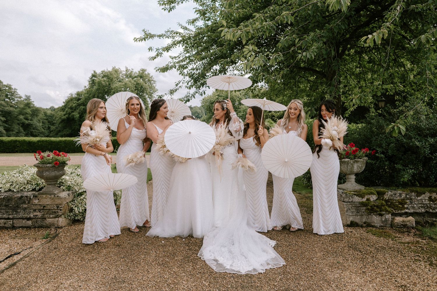 A bridal party in white dresses holding white parasols poses together on a gravel path near stone walls and trees.