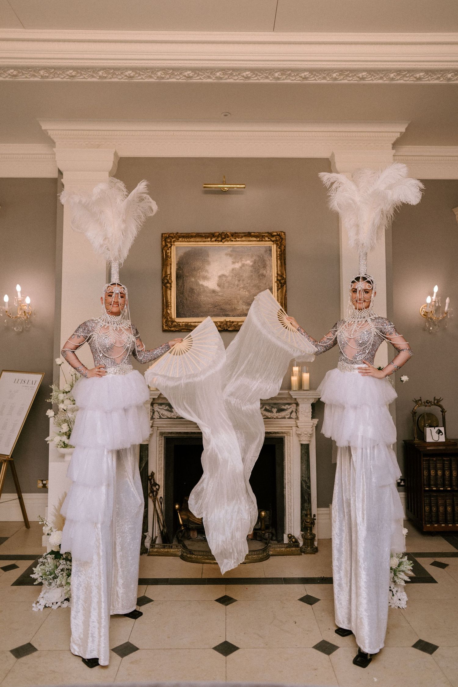 Three figures in white vintage-style performance costumes with tall feather headdresses pose elegantly in a classic interior setting.