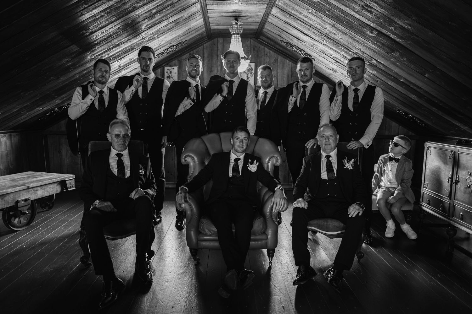 A black and white wedding photo showing a group of formally dressed groomsmen in suits arranged in two rows.