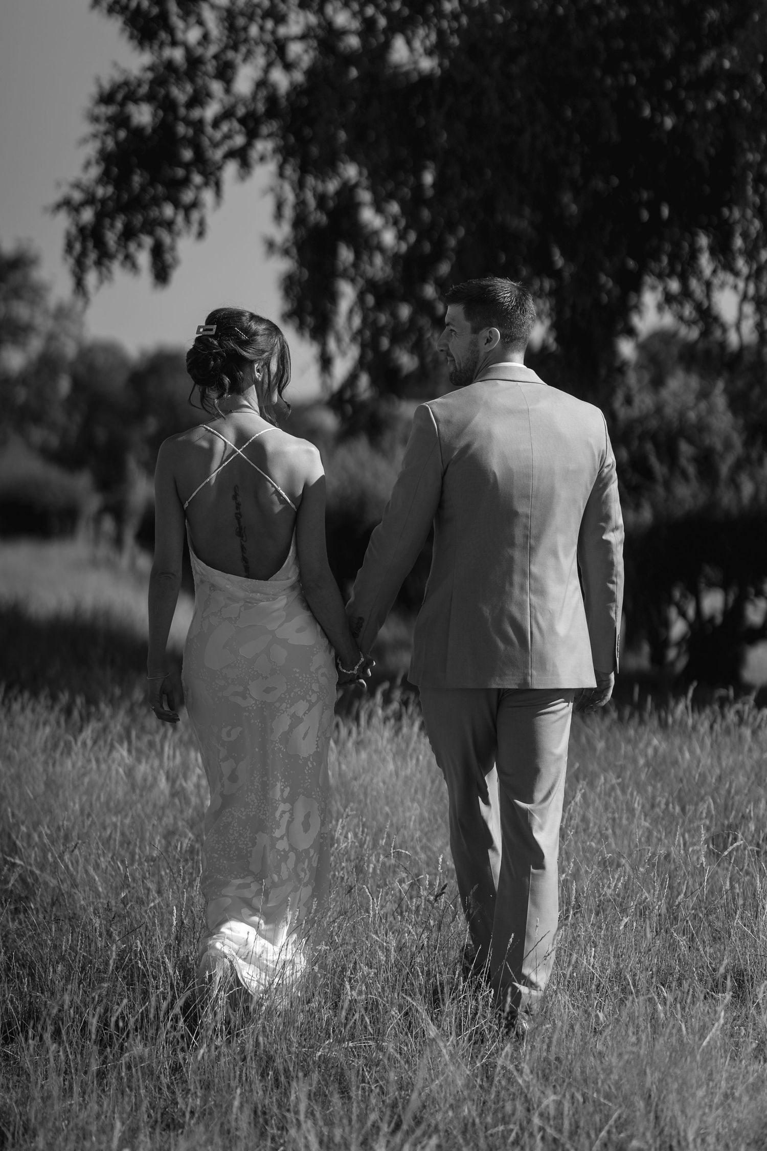 Black and white photo of a bride and groom holding hands walking through a grassy field toward trees.