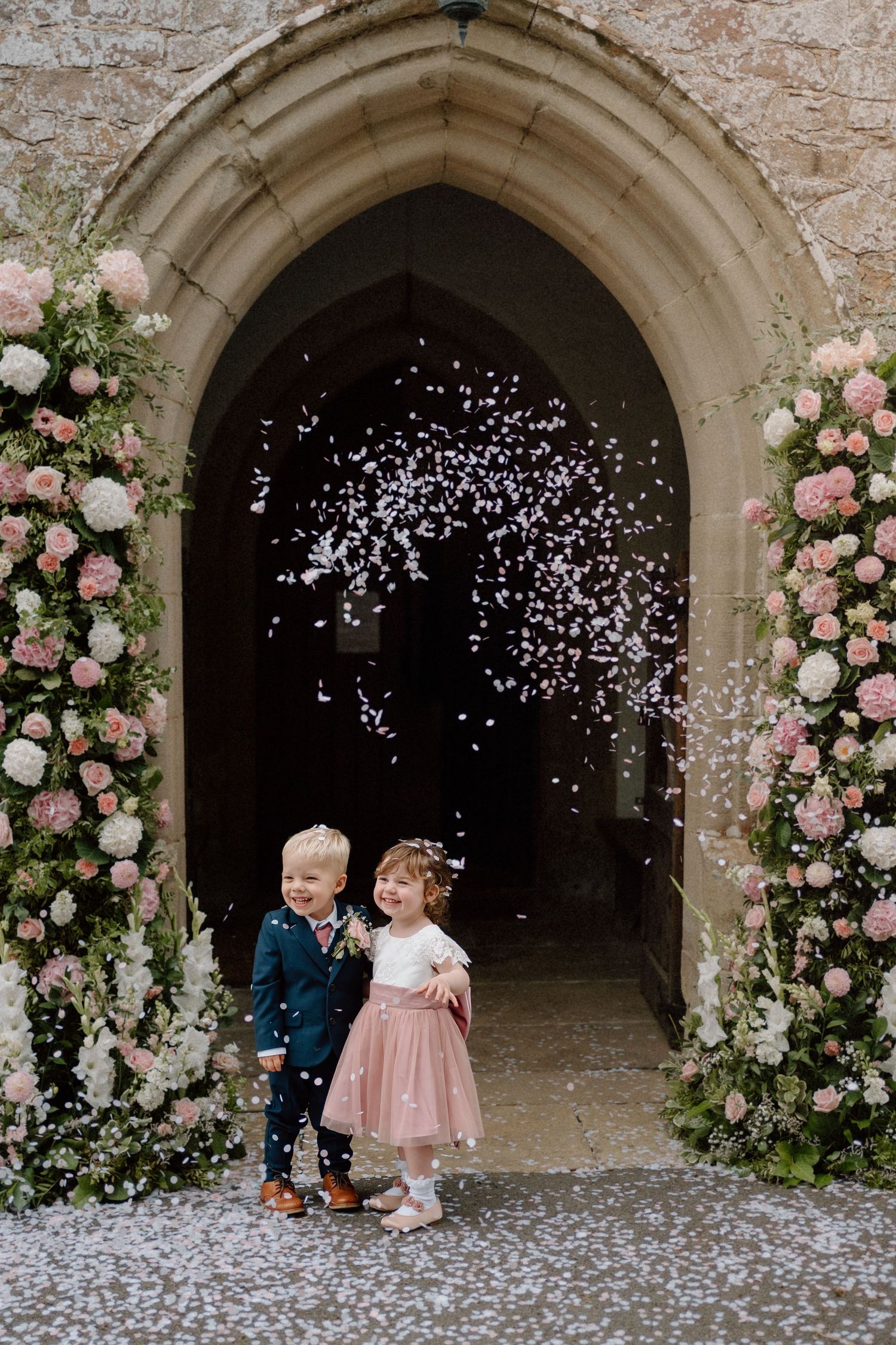 Two children stand beneath a decorative stone archway adorned with pink roses and white flowers while confetti falls around them.