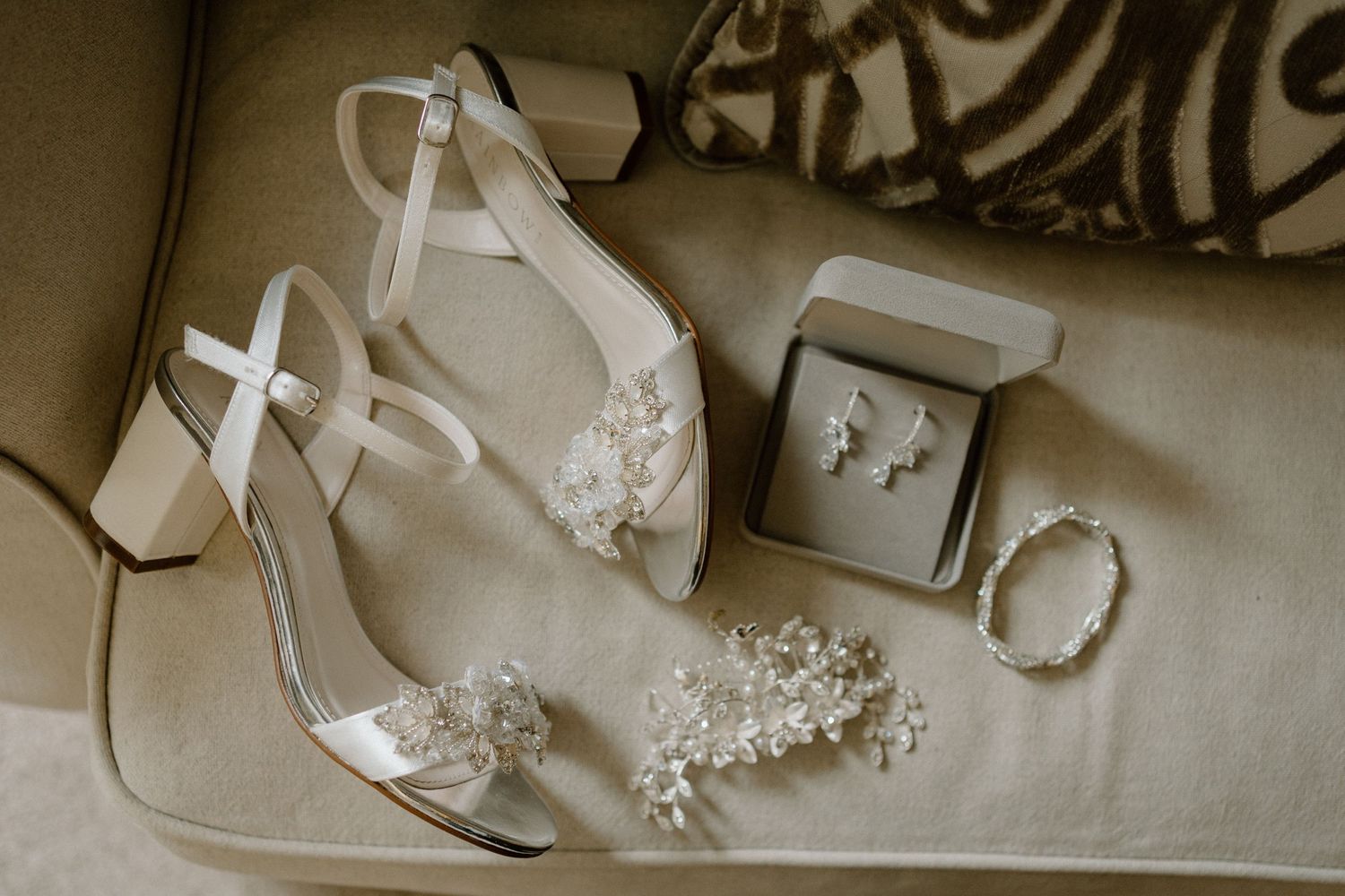 Wedding accessories including white heels with pearl details, delicate jewelry and earrings arranged on beige fabric at Dodford Manor.
