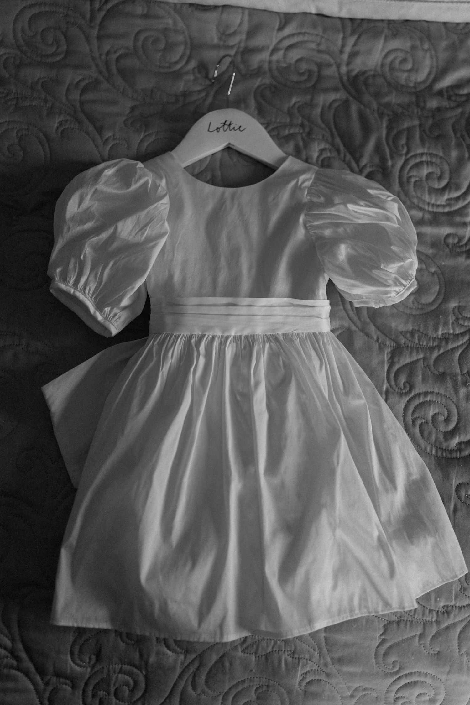 White puff-sleeve flower girl dress with sash displayed on hanger against textured background at Dodford Manor.