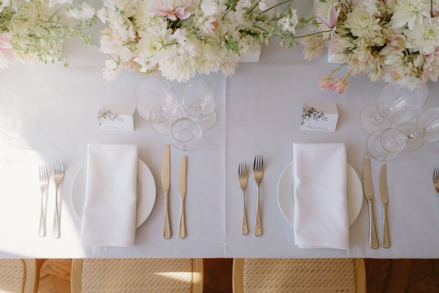 Elegant wedding table setting with gold cutlery, white napkins, and blush floral arrangements overhead.
