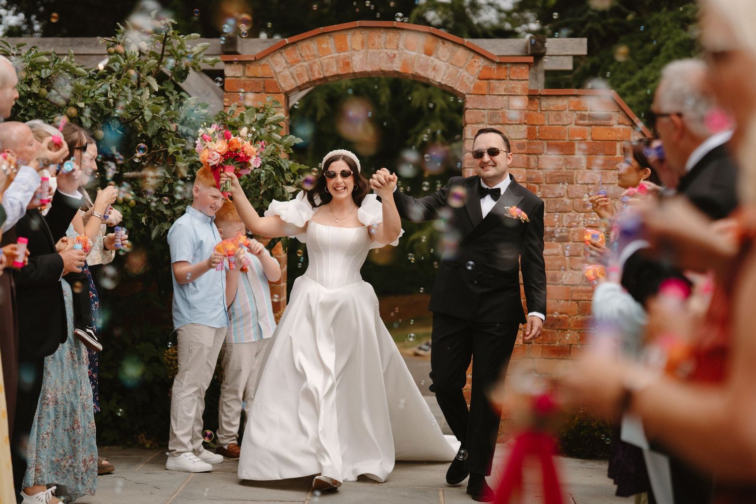 Bride and groom walk through bubble send-off under brick arch surrounded by cheering guests at outdoor wedding.