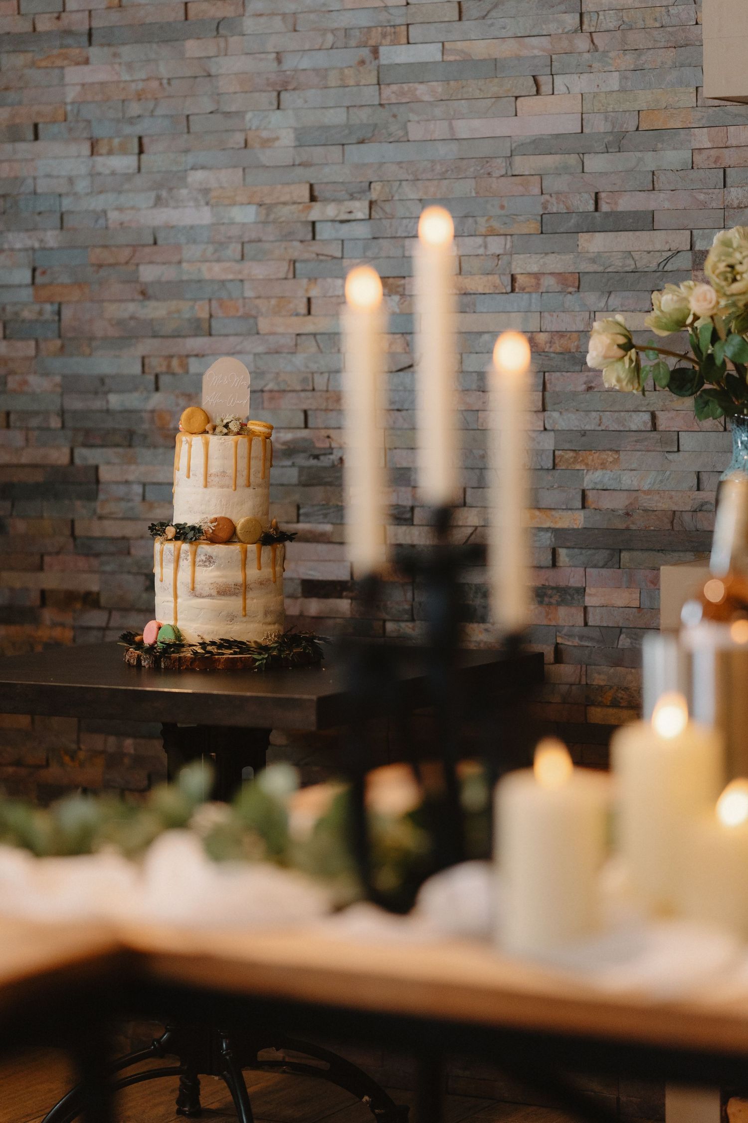 Elegant two-tier semi-naked wedding cake with caramel drip, displayed beside lit taper candles against a stone wall.