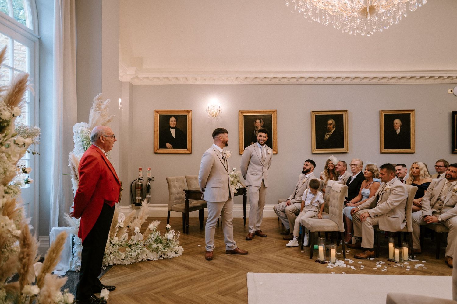 Wedding ceremony in an elegant white room with framed portraits, floral arrangements and a crystal chandelier.