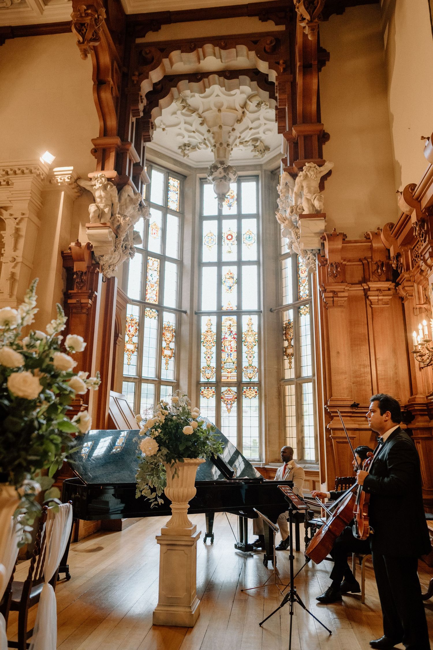 Musicians perform by tall ornate windows in an elegant wood-panelled room with floral arrangements at Harlaxton Manor.