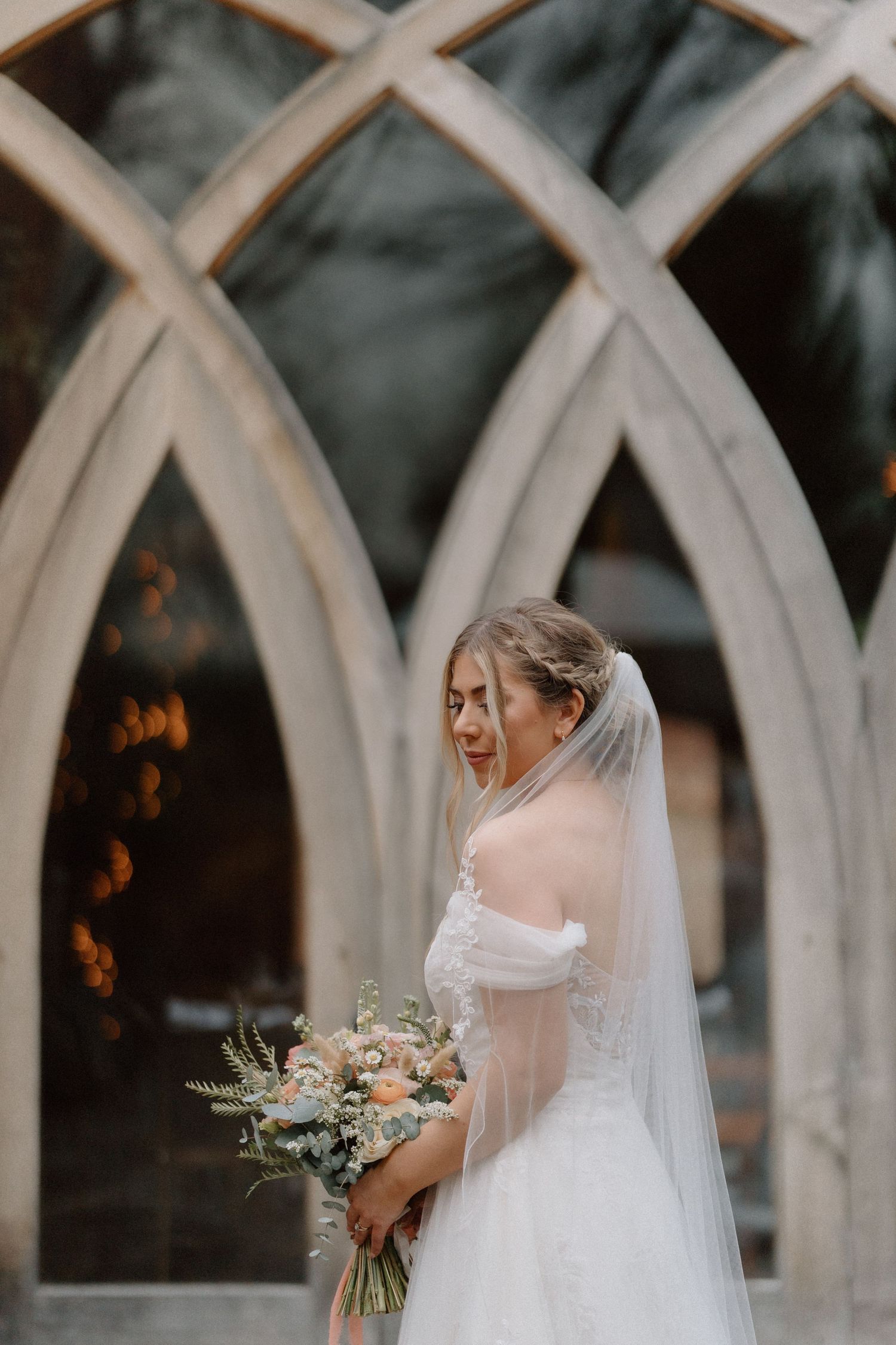 Ethereal bridal portrait through gothic arched windows with a soft bouquet and flowing veil at Shustoke Barn.