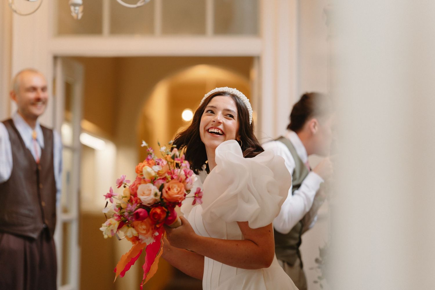 Joyful bride laughing while holding a vibrant colorful bouquet of pink and orange flowers in an elegant venue.
