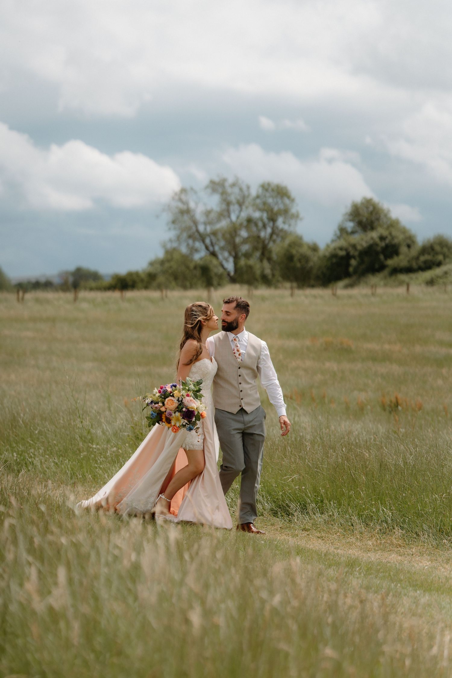 A bride and groom walk hand-in-hand through a lush green meadow, she holds a colorful bouquet in her flowing gown.